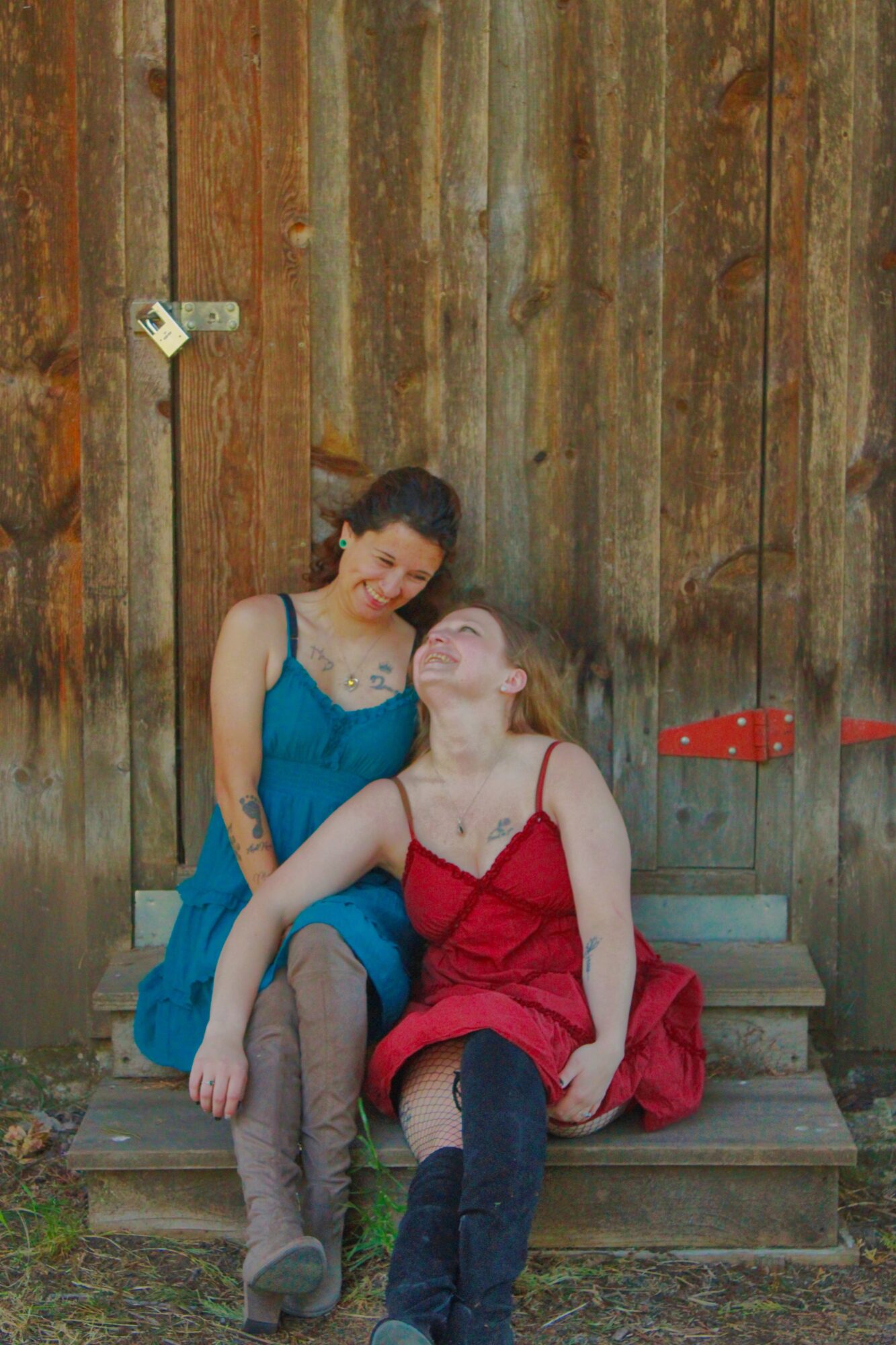 Two women sitting on a wooden bench in front of a wooden wall, one in a blue dress and the other in a red dress, smiling and leaning towards each other.