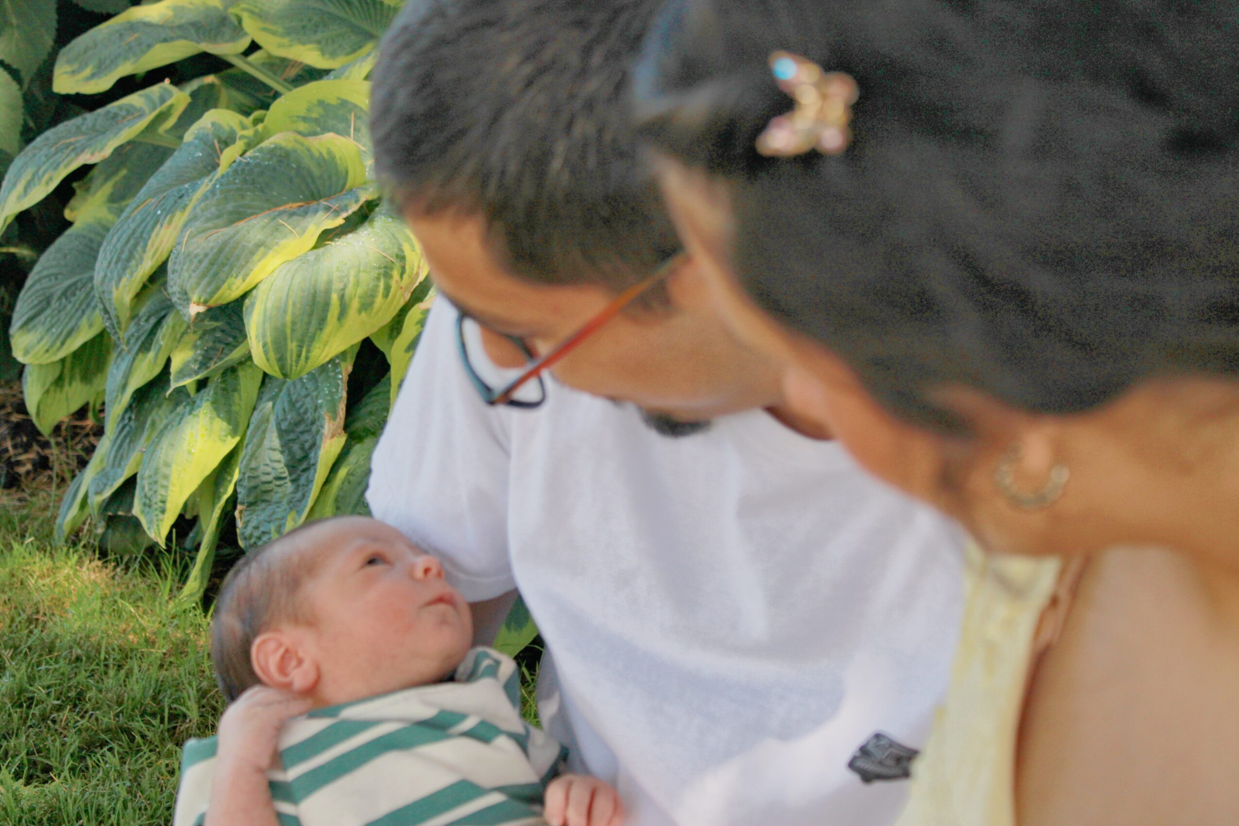 Two adults looking at a baby outdoors with green foliage in the background.