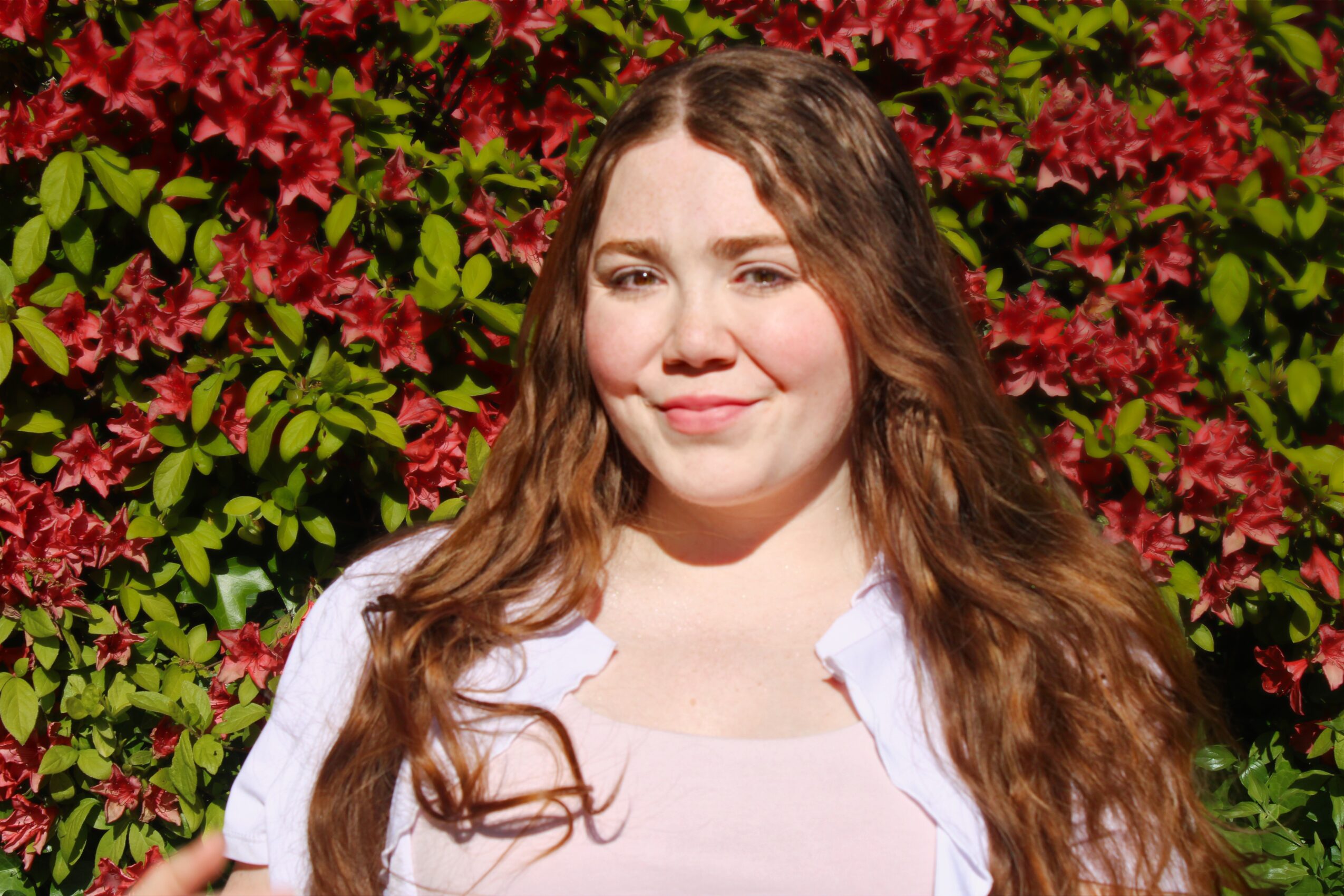 Young woman with long wavy hair standing in front of pink and green flowering bushes, smiling softly.