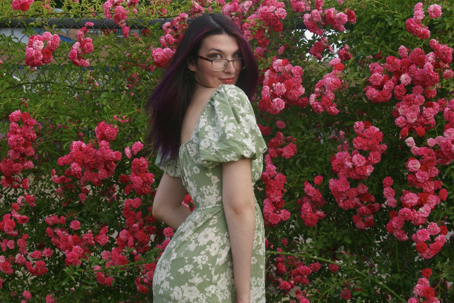 Young woman with glasses and long dark hair in a floral dress standing among pink roses in a garden.