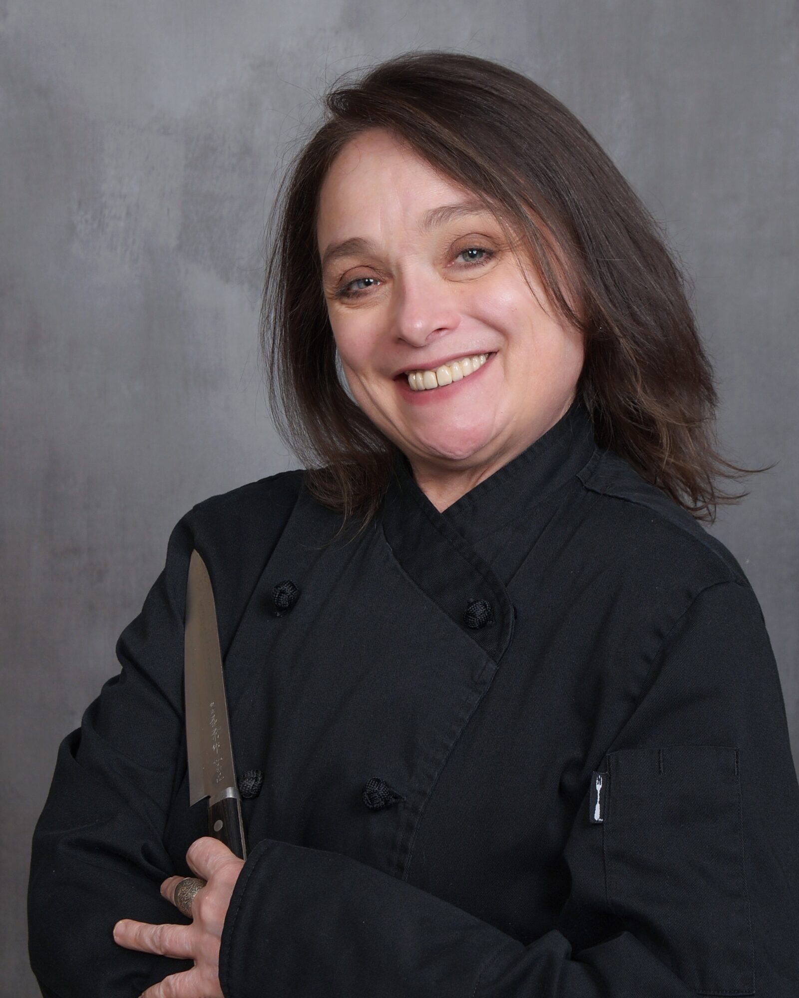 Woman with shoulder-length brown hair smiling, wearing a black chef's coat, holding a knife, standing against a gray background.