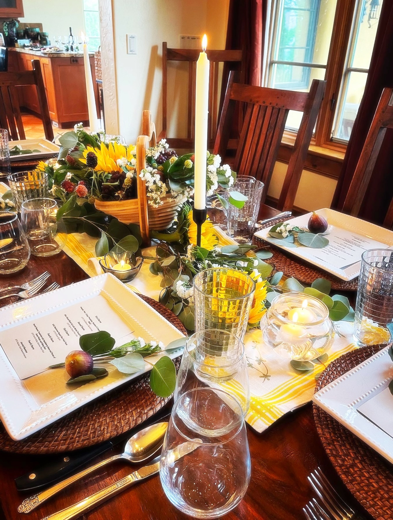 Dining table with floral centerpiece, candles, glasses, plates, and cutlery, set near windows with natural light.