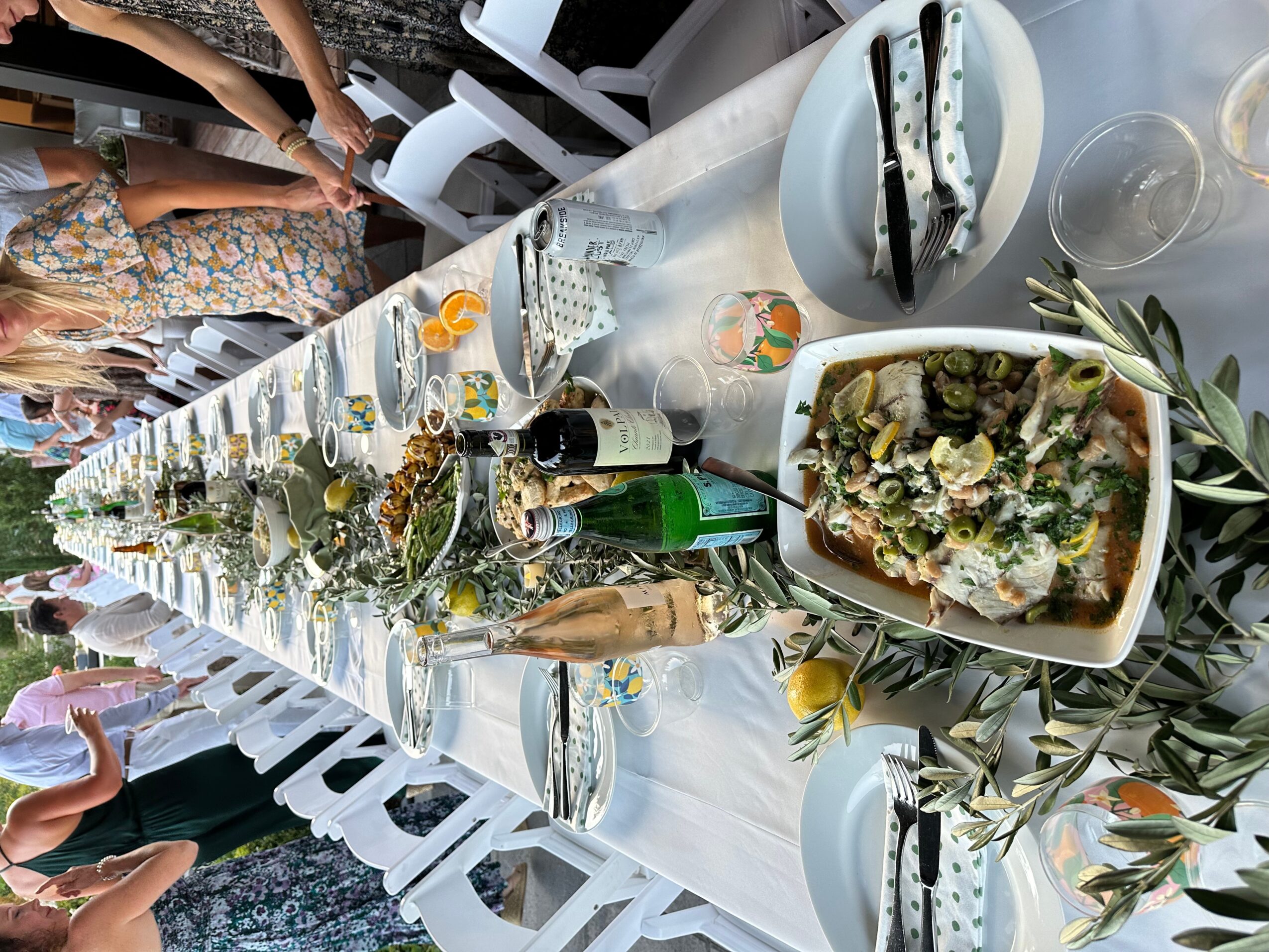 Long table set with plates, glasses, and food, with people standing around outdoors, decorated with greenery and lemons.
