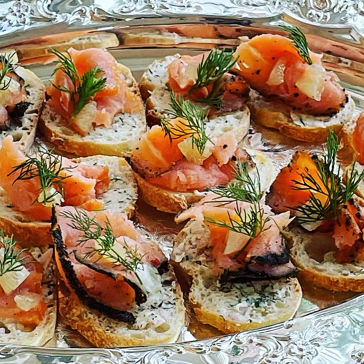 Assorted smoked fish and cream cheese on bread slices garnished with herbs on a silver platter.