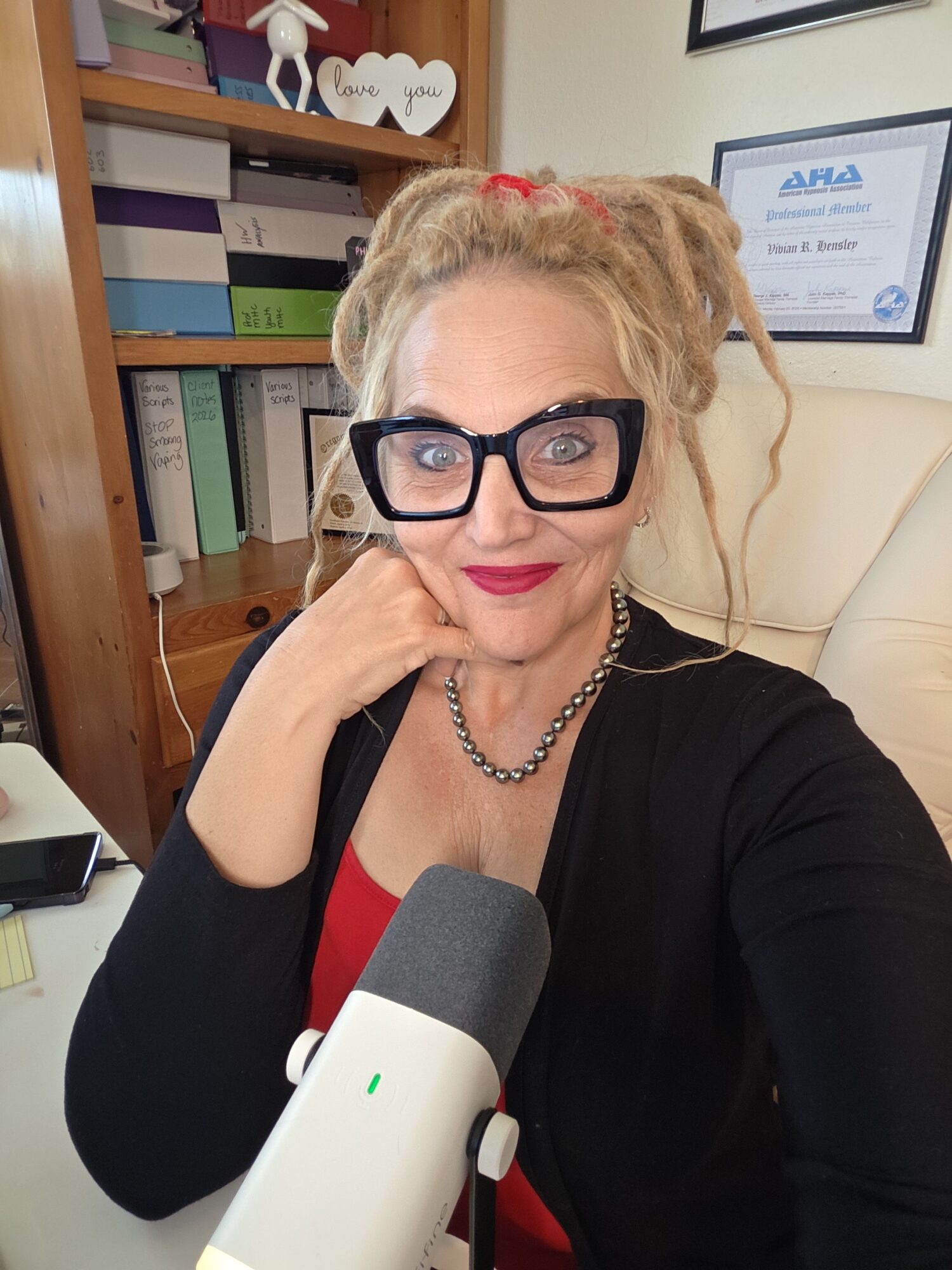 Woman with glasses and blonde hair, wearing a black blazer, sitting in an office with bookshelves behind her.