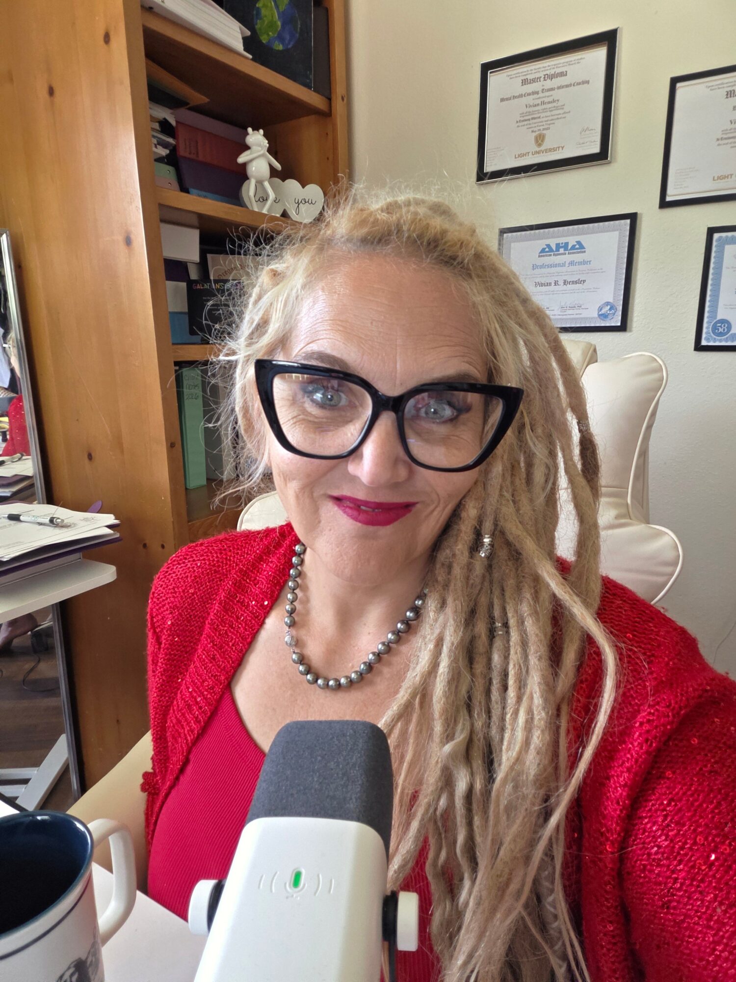 Woman with glasses and blonde dreadlocks wearing a red sweater, sitting at a desk with books and framed certificates on the wall.