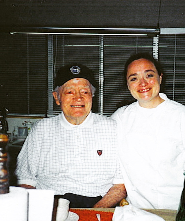 Two people smiling indoors, one older man with gray hair and a woman with dark hair, both wearing white shirts.