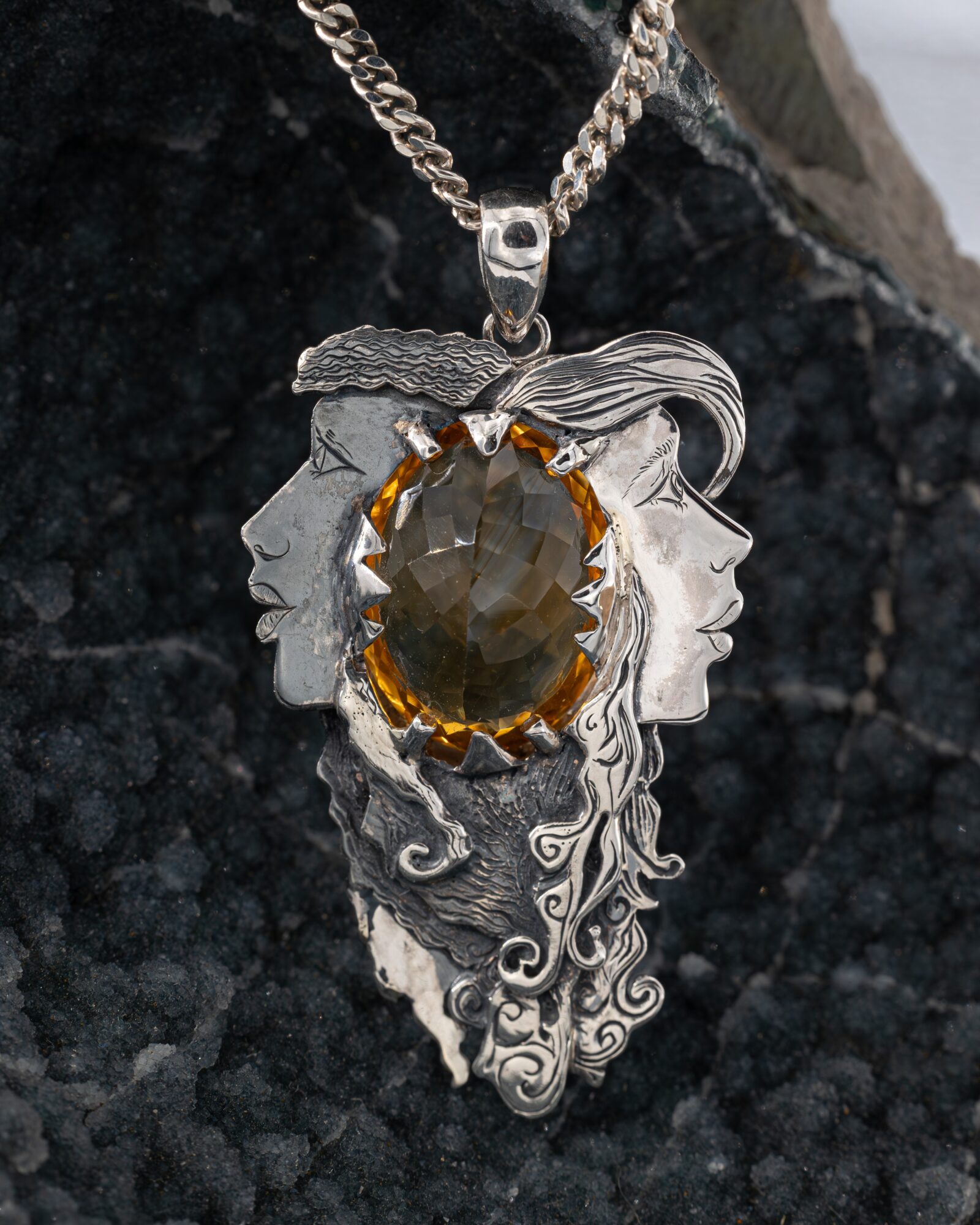 Pendant with two faces, one male and one female, surrounding a large oval gemstone, on a chain, against a dark background.