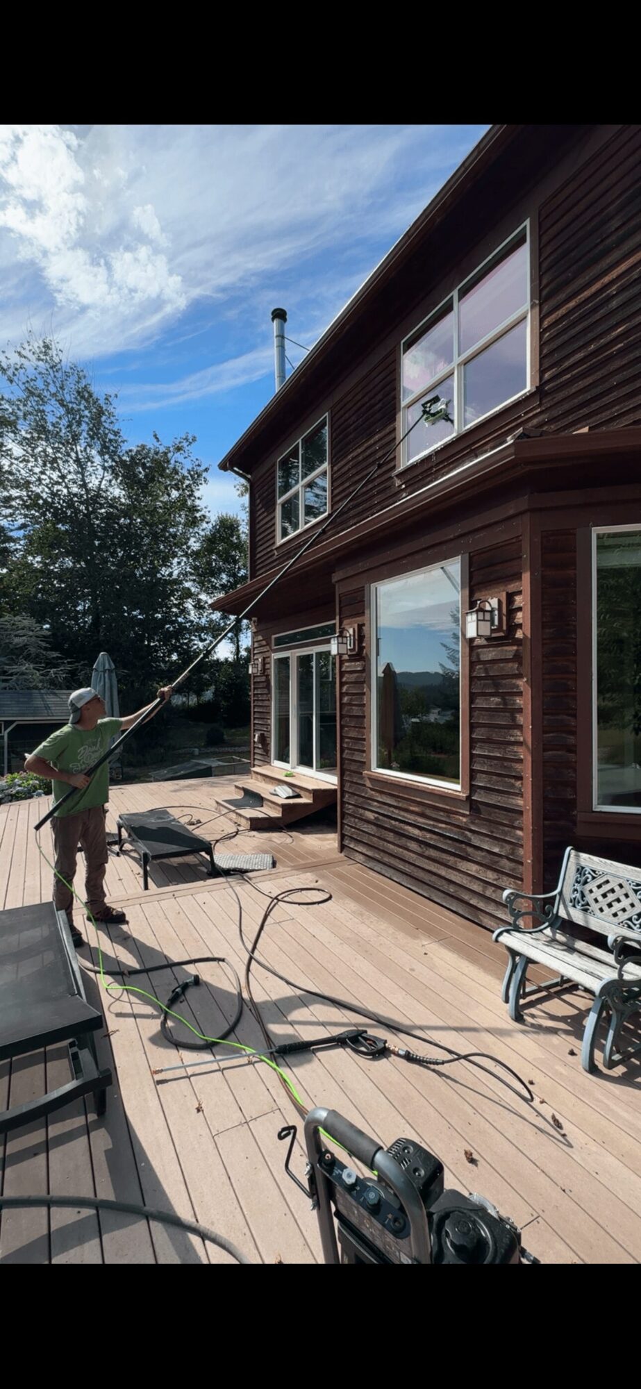 Person pressure washing wooden deck outside house with large windows, trees, and blue sky in background.
