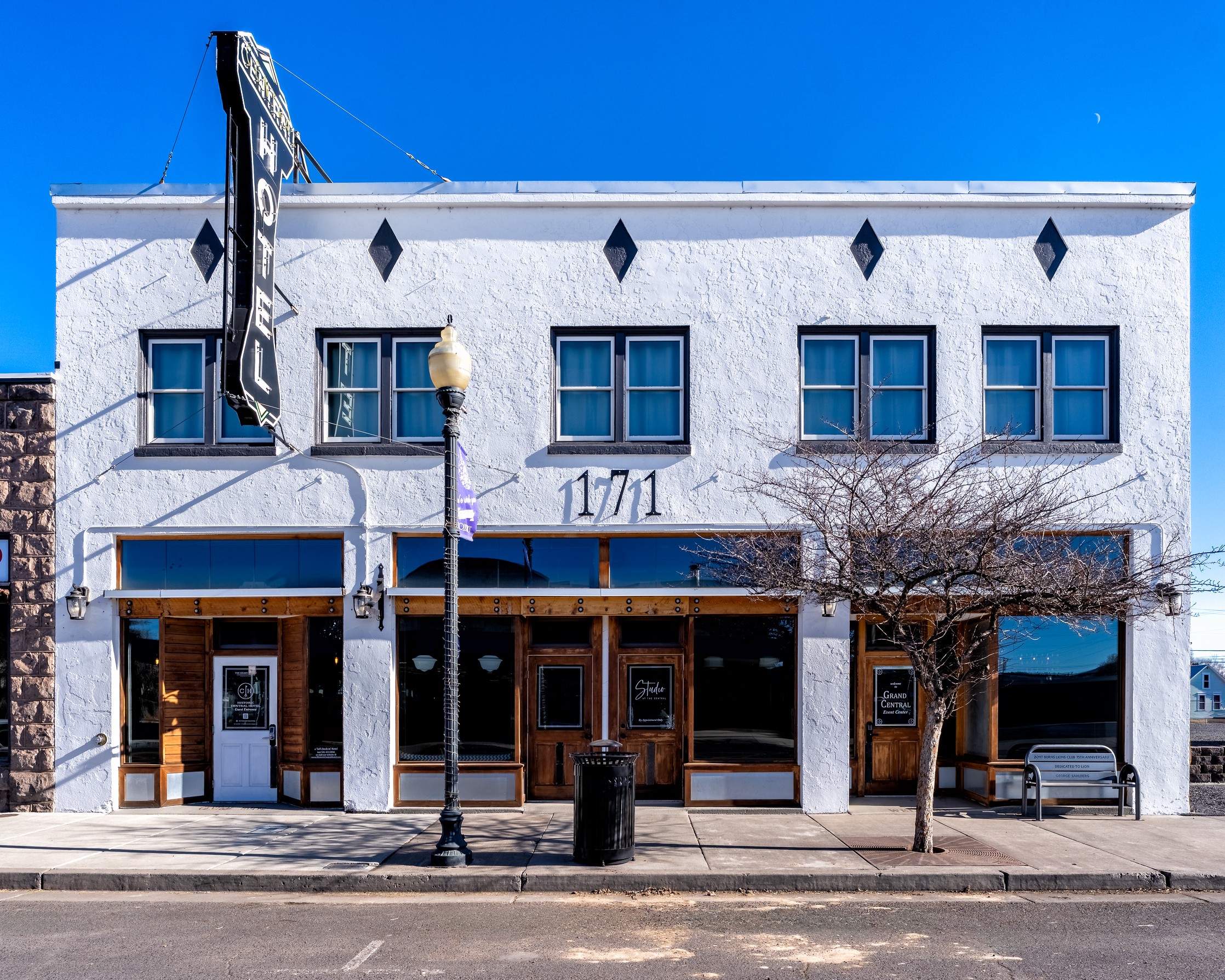 White two-story building with a sign, multiple windows, and a sidewalk with trees and a lamppost.