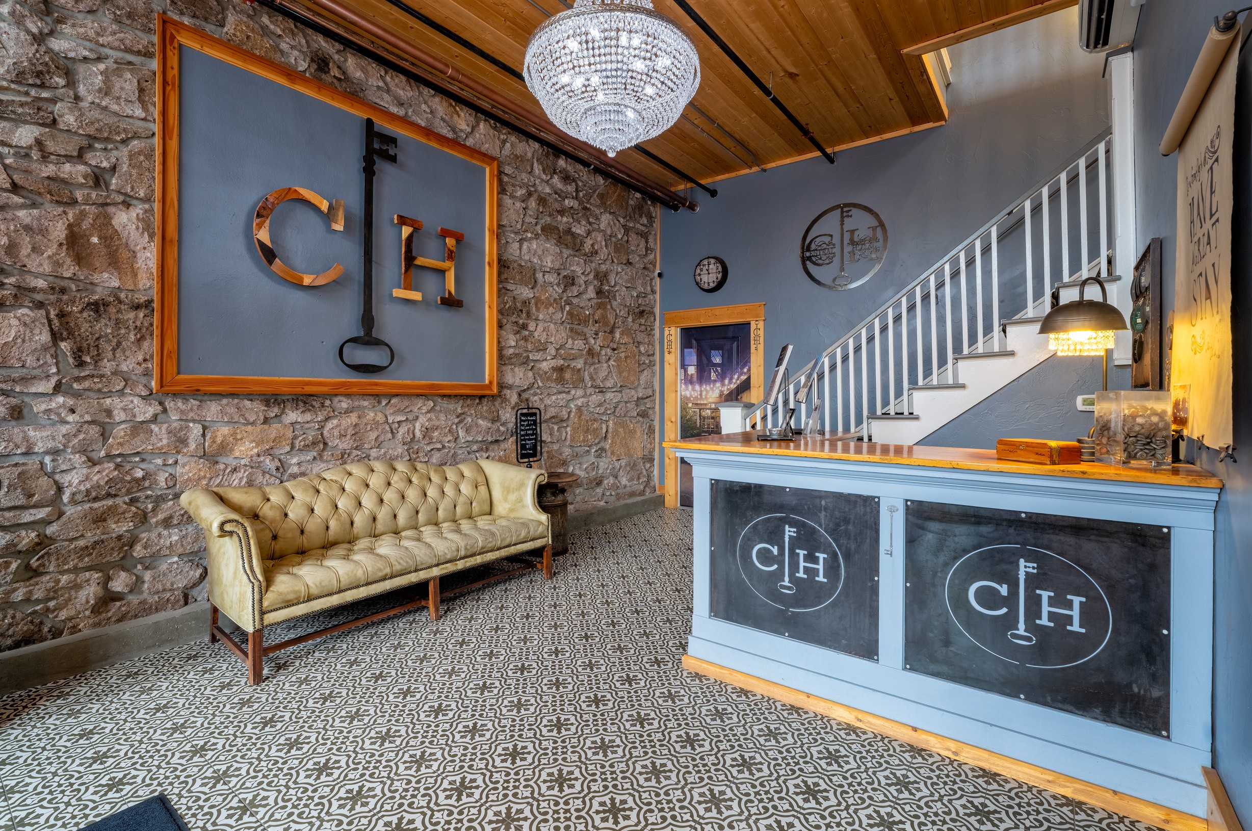 Reception area with a vintage sofa, brick wall, CH logo artwork, and a blue front desk with CH logo, staircase nearby.