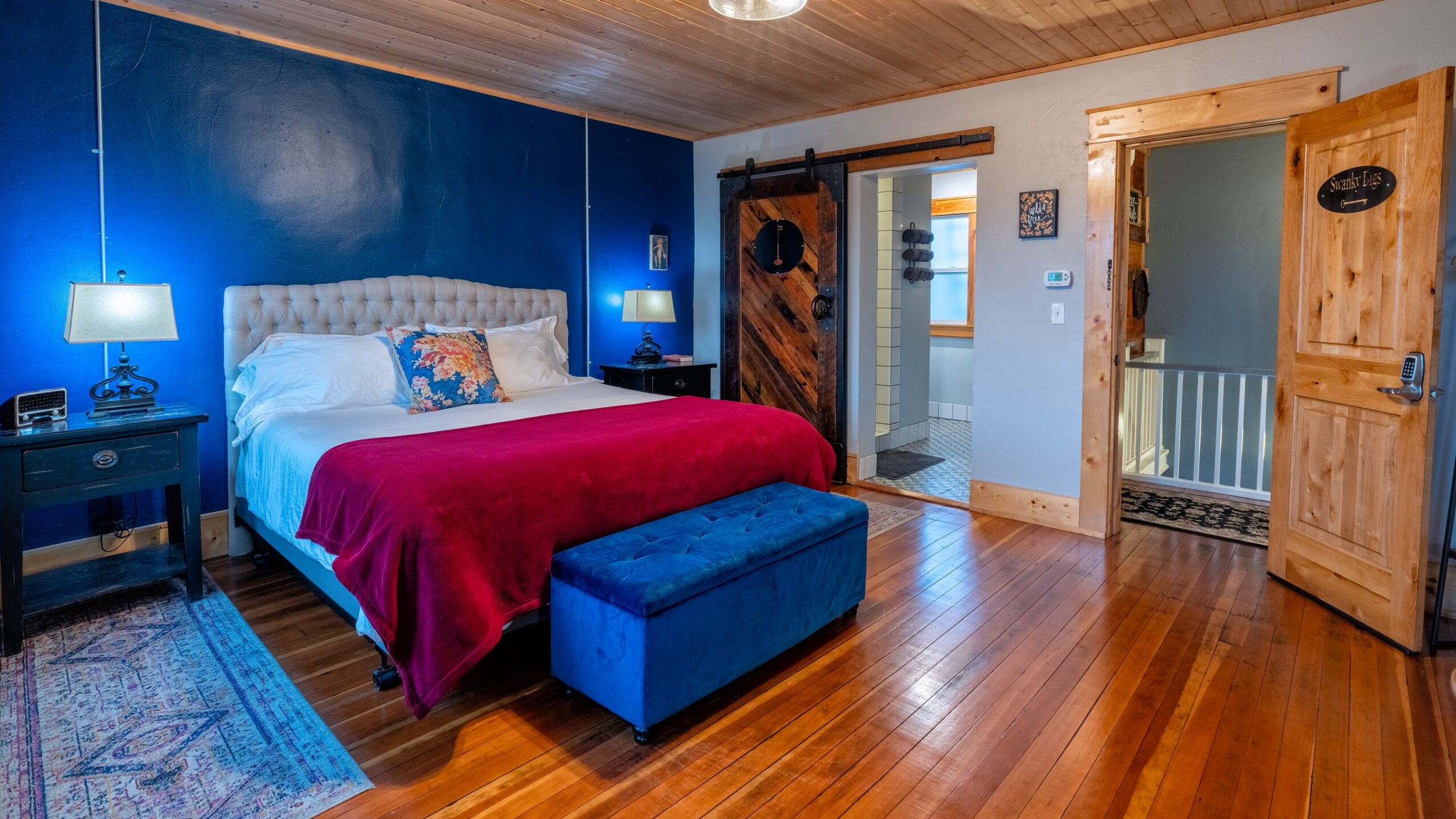 Bedroom with blue accent wall, bed with white headboard, red blanket, blue ottoman, wooden floors, and two doors.