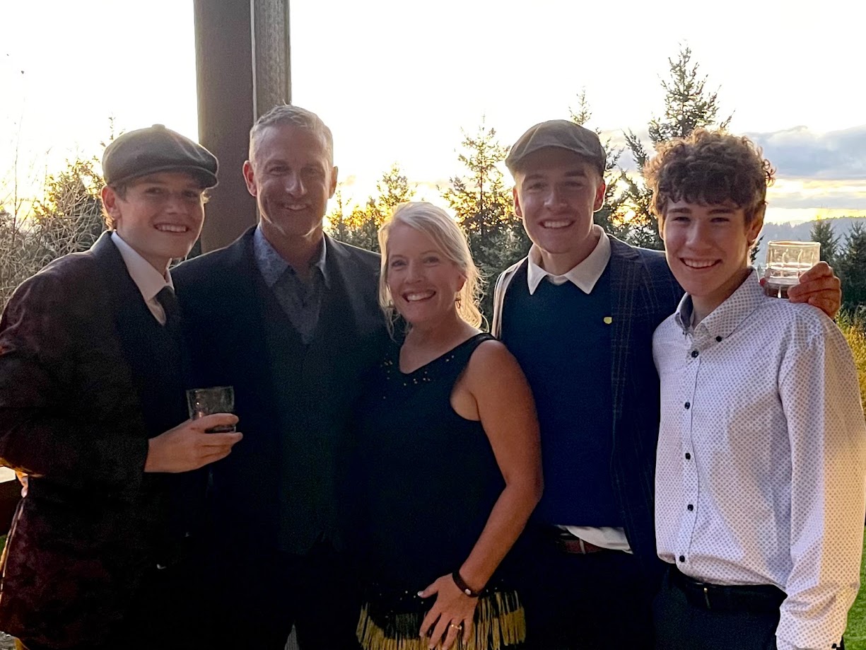 Group of five people smiling outdoors, some holding drinks, with trees and sky in background.