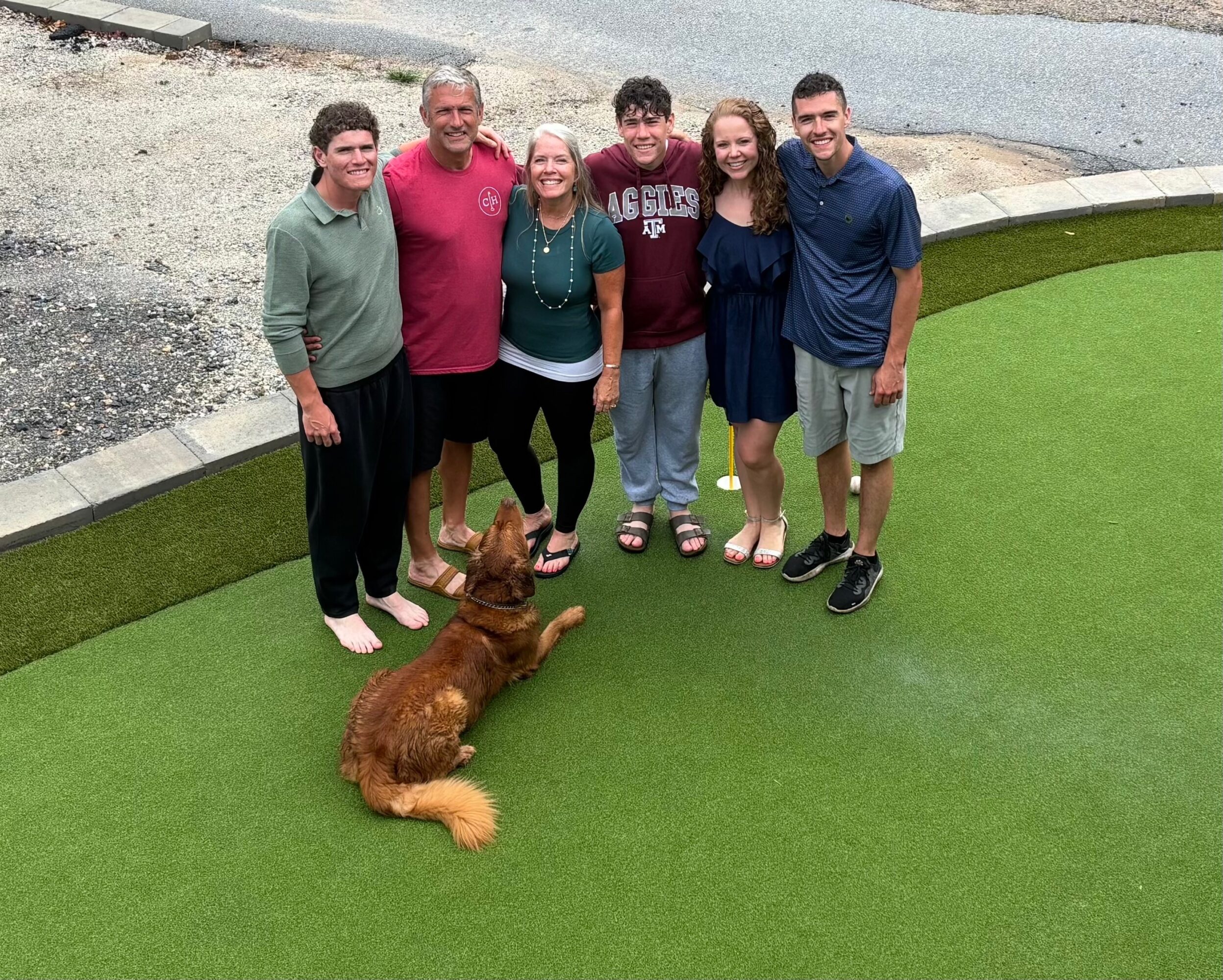Group of six people standing outdoors on green artificial grass with a dog lying in front, smiling at the camera.