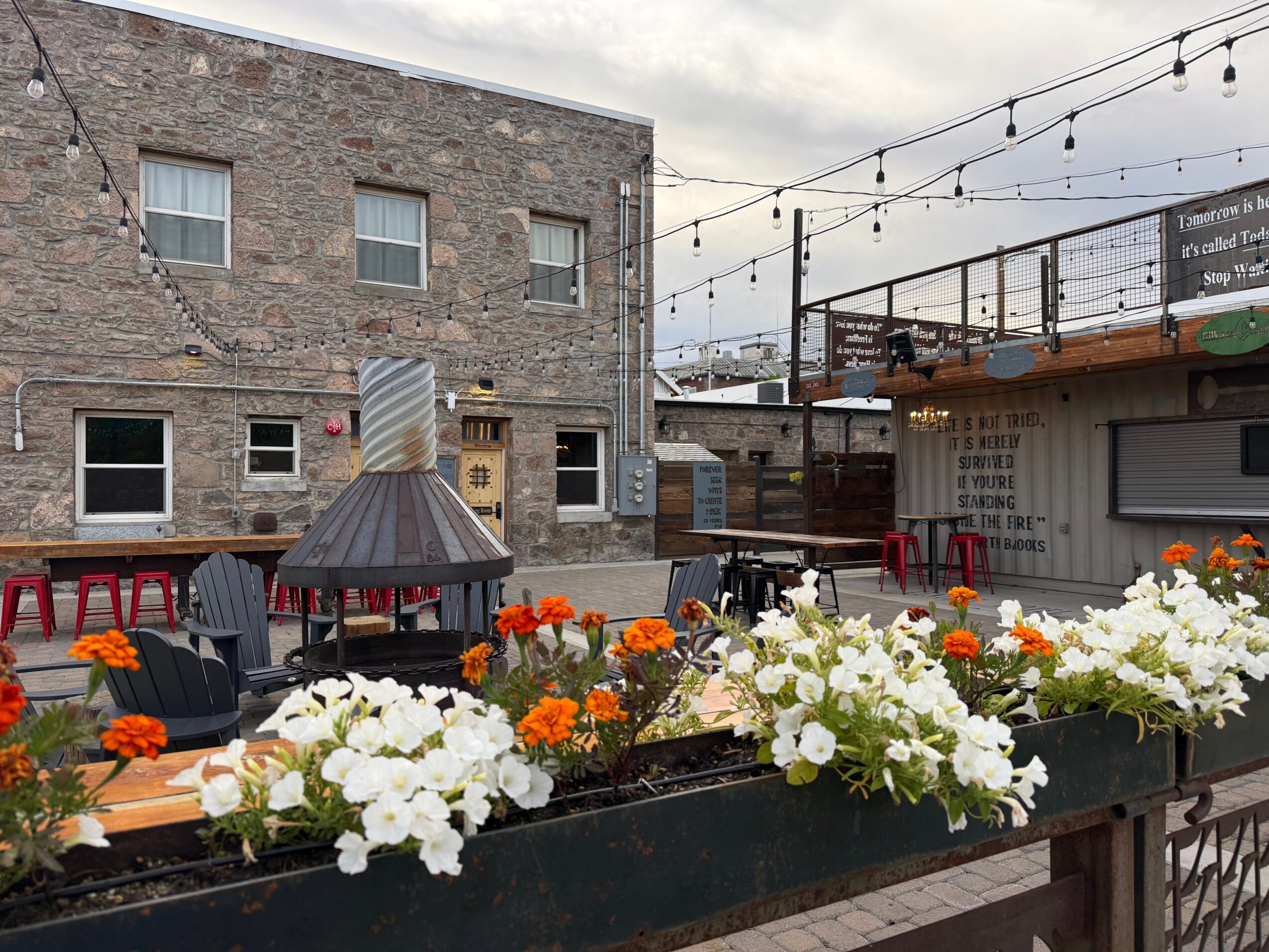 Outdoor patio with flower boxes, string lights, and seating area against a brick building background.