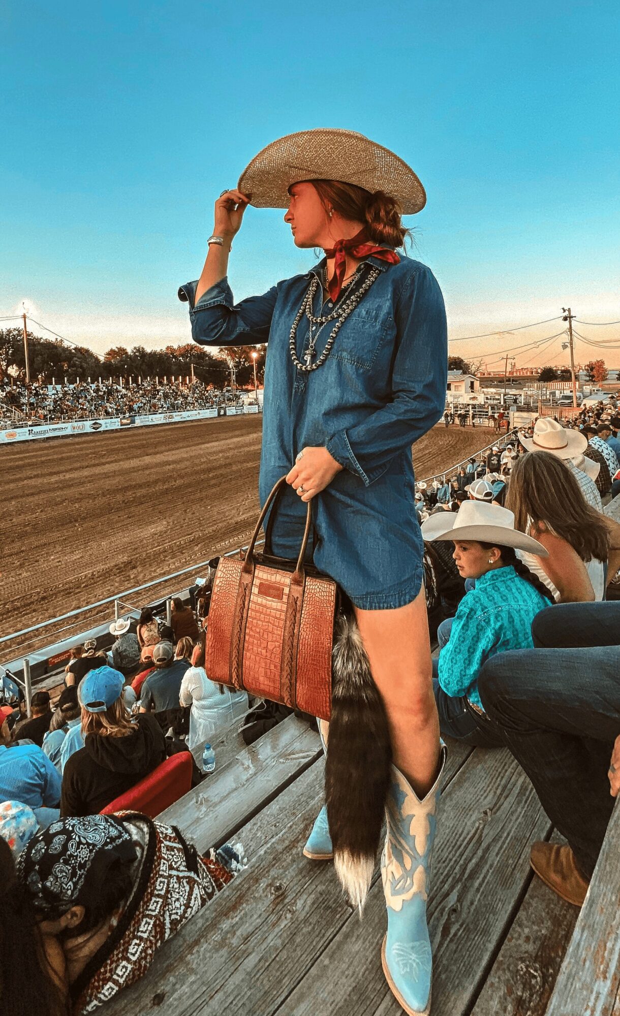 Woman in denim outfit with cowboy hat and boots, holding a bag, at a rodeo event with spectators in background.