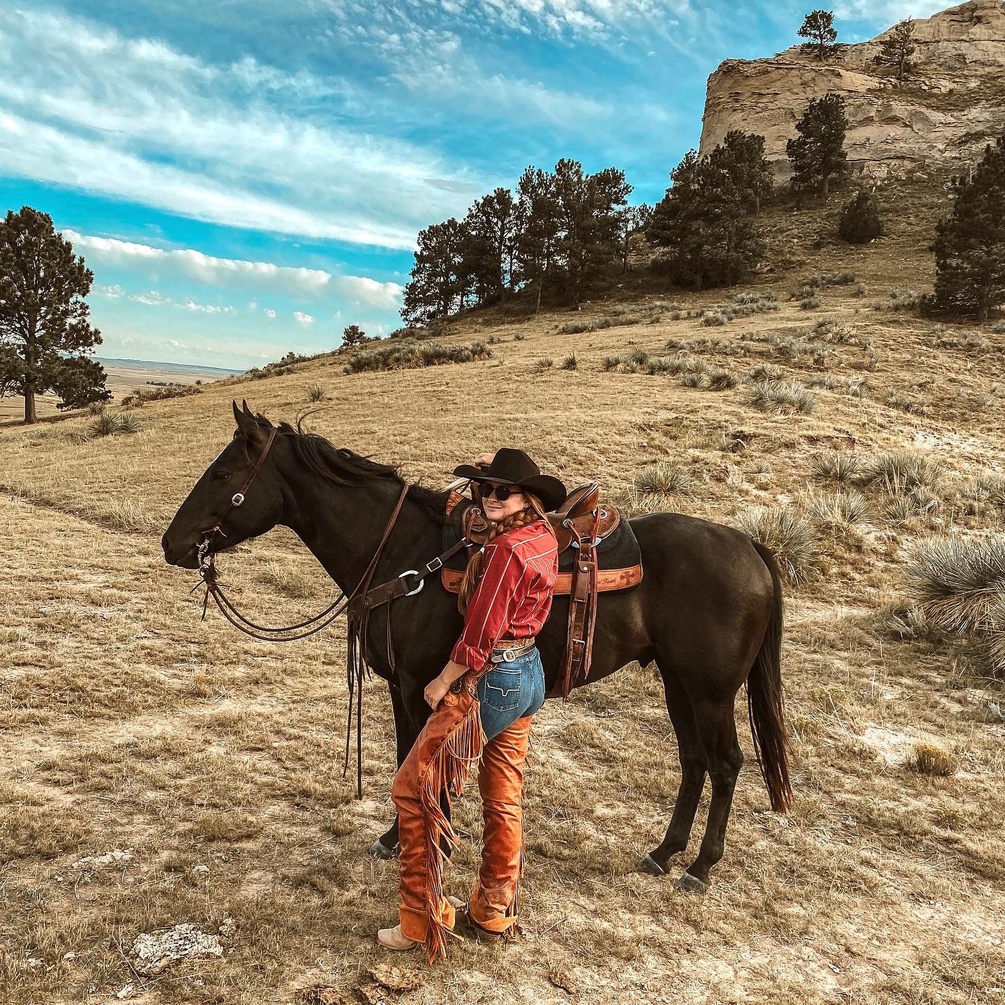 Person in cowboy attire standing next to a black horse in a dry, open landscape with trees and a rocky hill, under a partly cloudy sky.