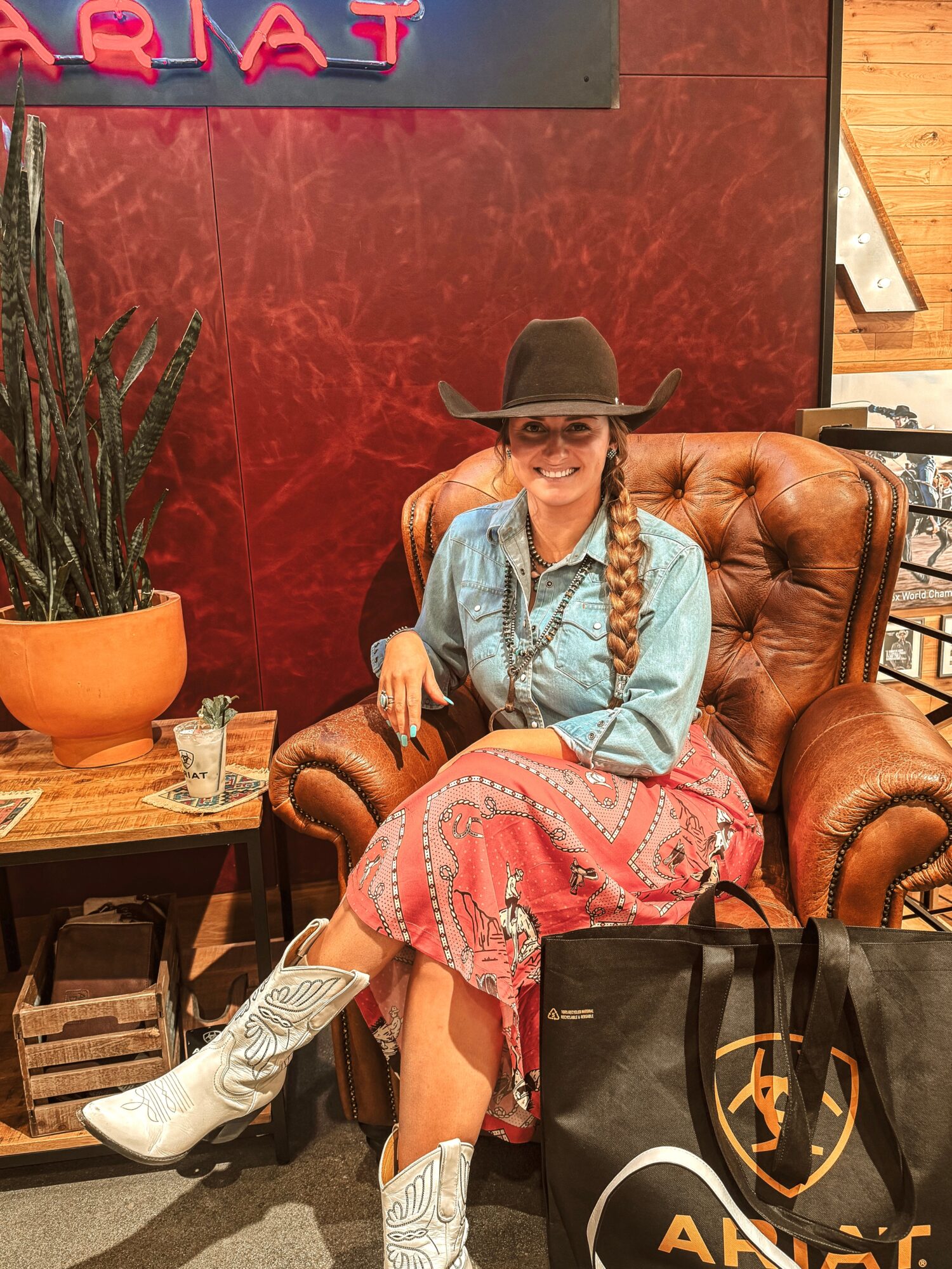 Young woman with long braided hair, wearing a cowboy hat, denim shirt, and pink skirt, sitting on a leather armchair.