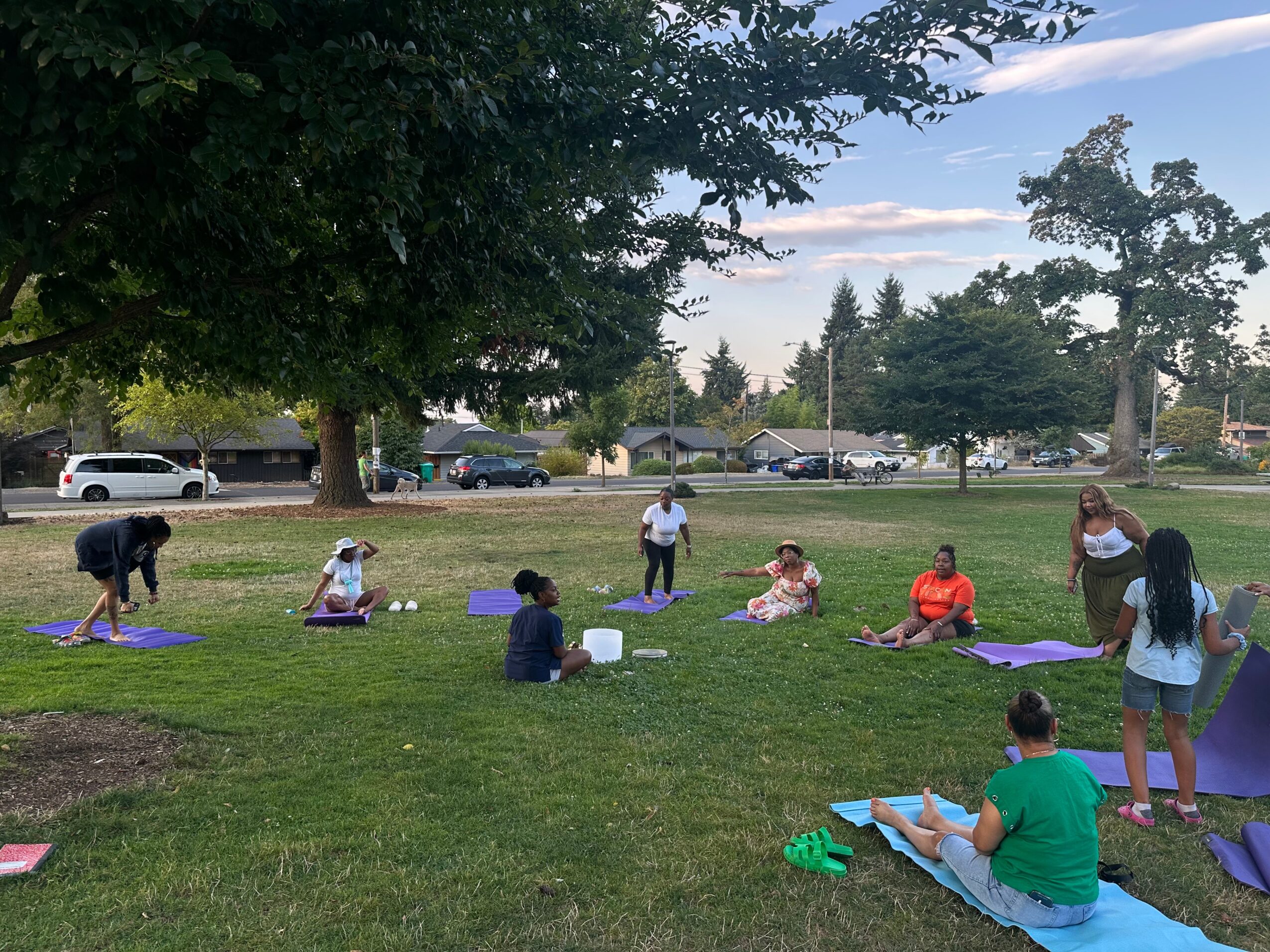 People practicing yoga outdoors on mats in a park with trees and cars in the background.