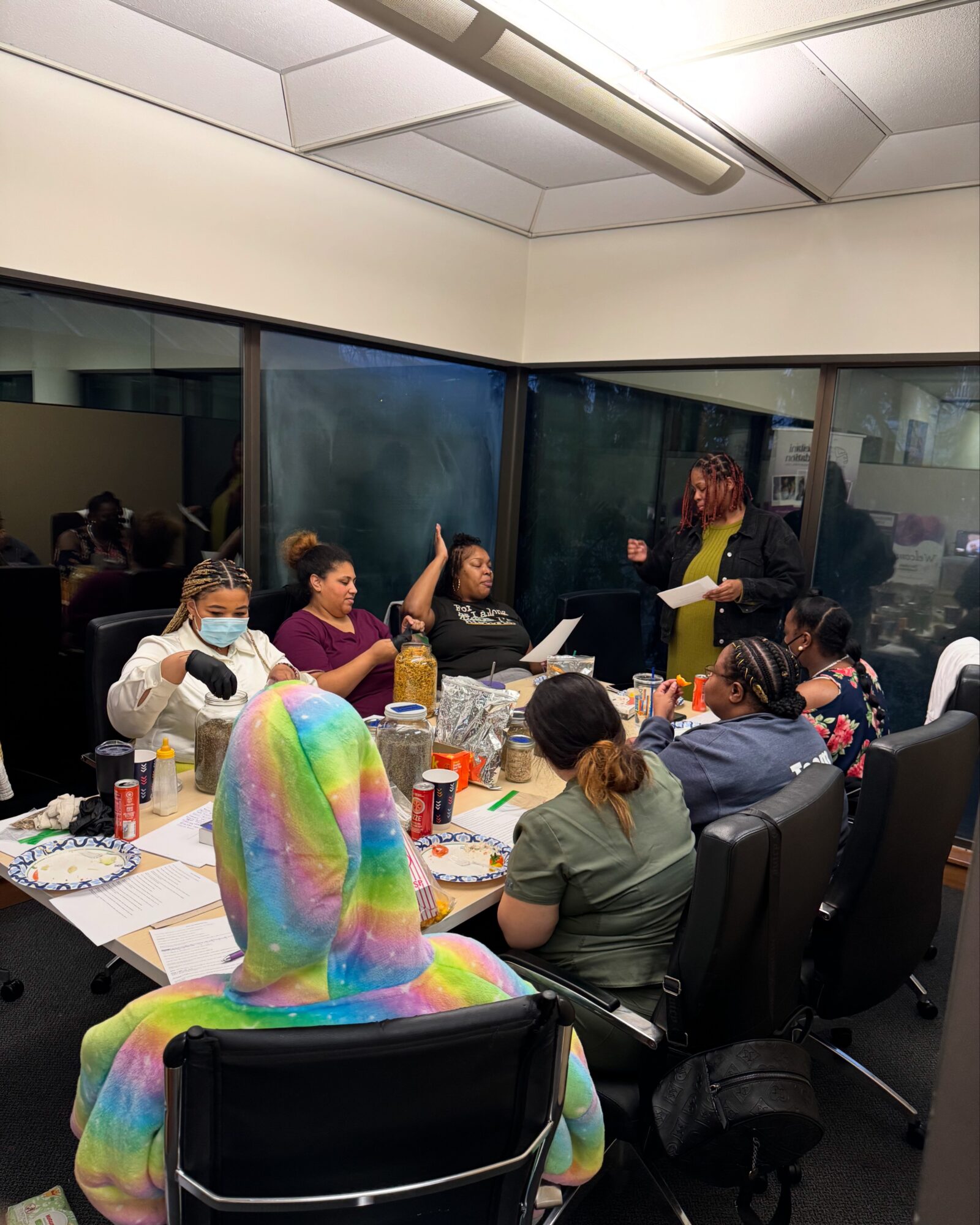 Group of people seated around a table in a conference room, some wearing masks, a woman standing and speaking, large windows in background.