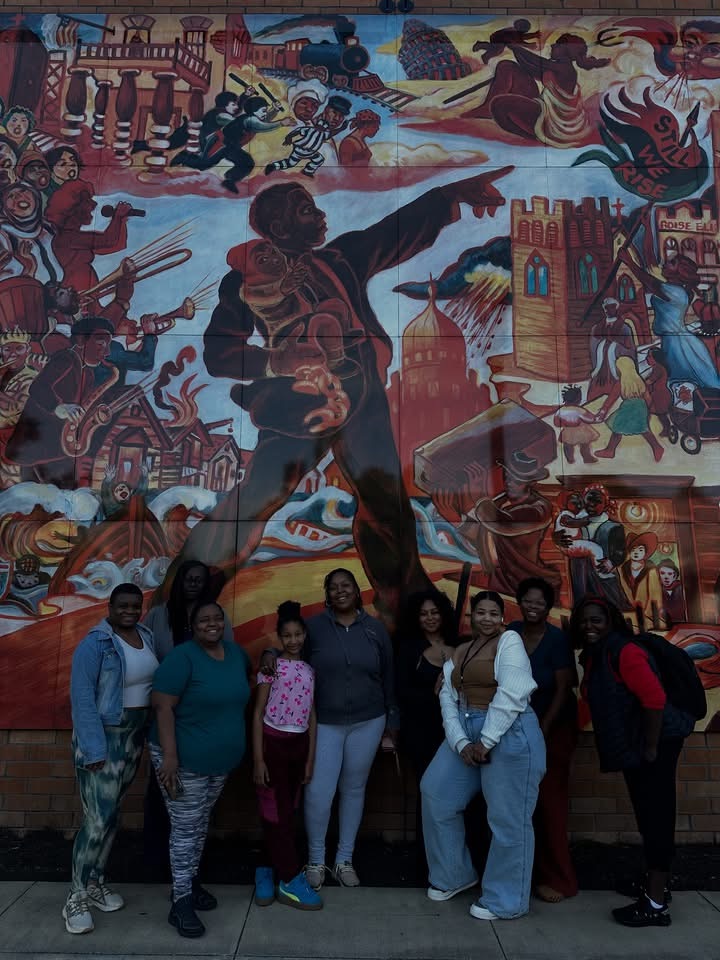 Group of eight people standing in front of a colorful mural depicting historical and cultural scenes.