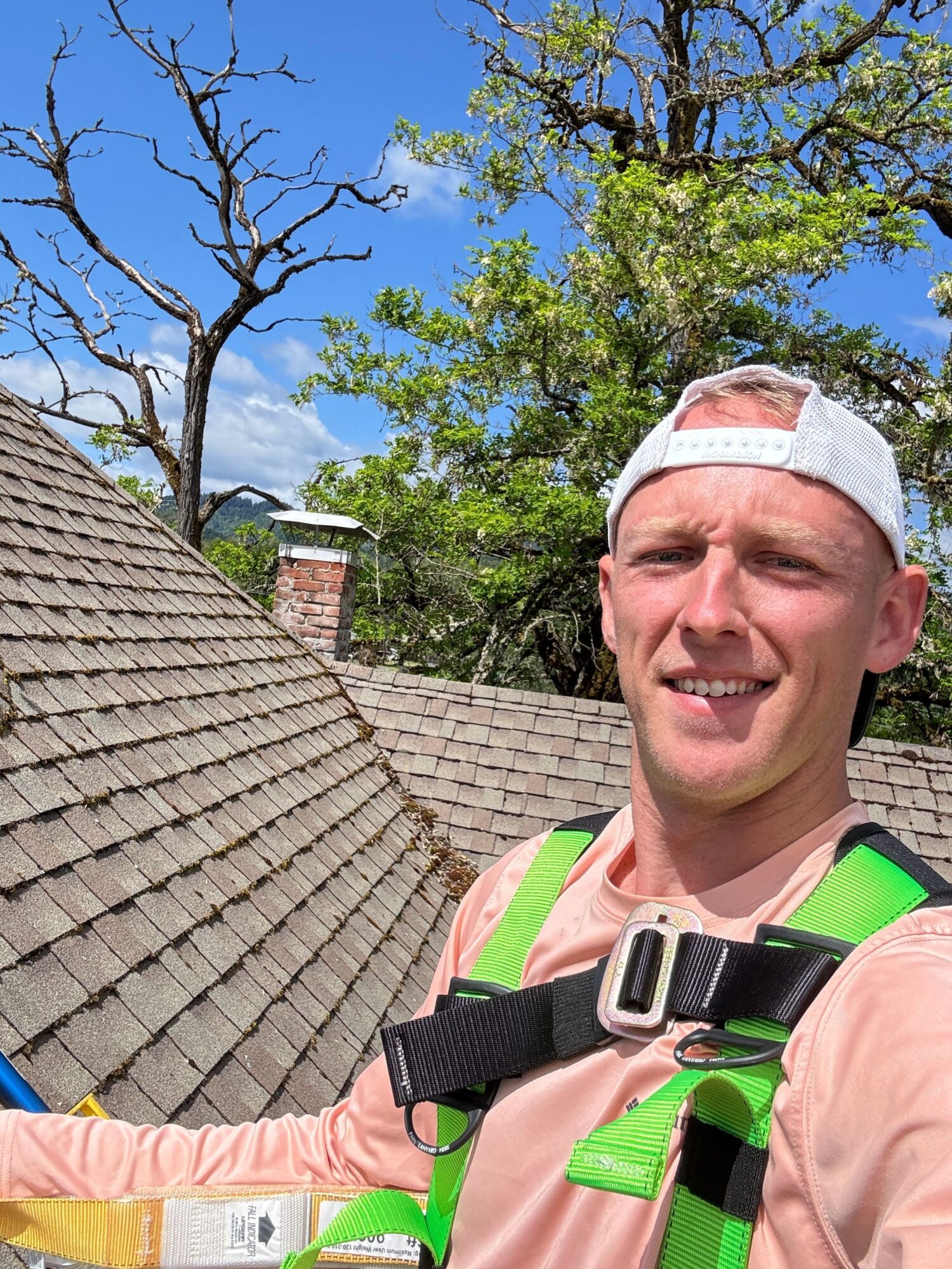 Person smiling outdoors with trees and blue sky, wearing a white cap and a pink shirt, carrying a backpack.