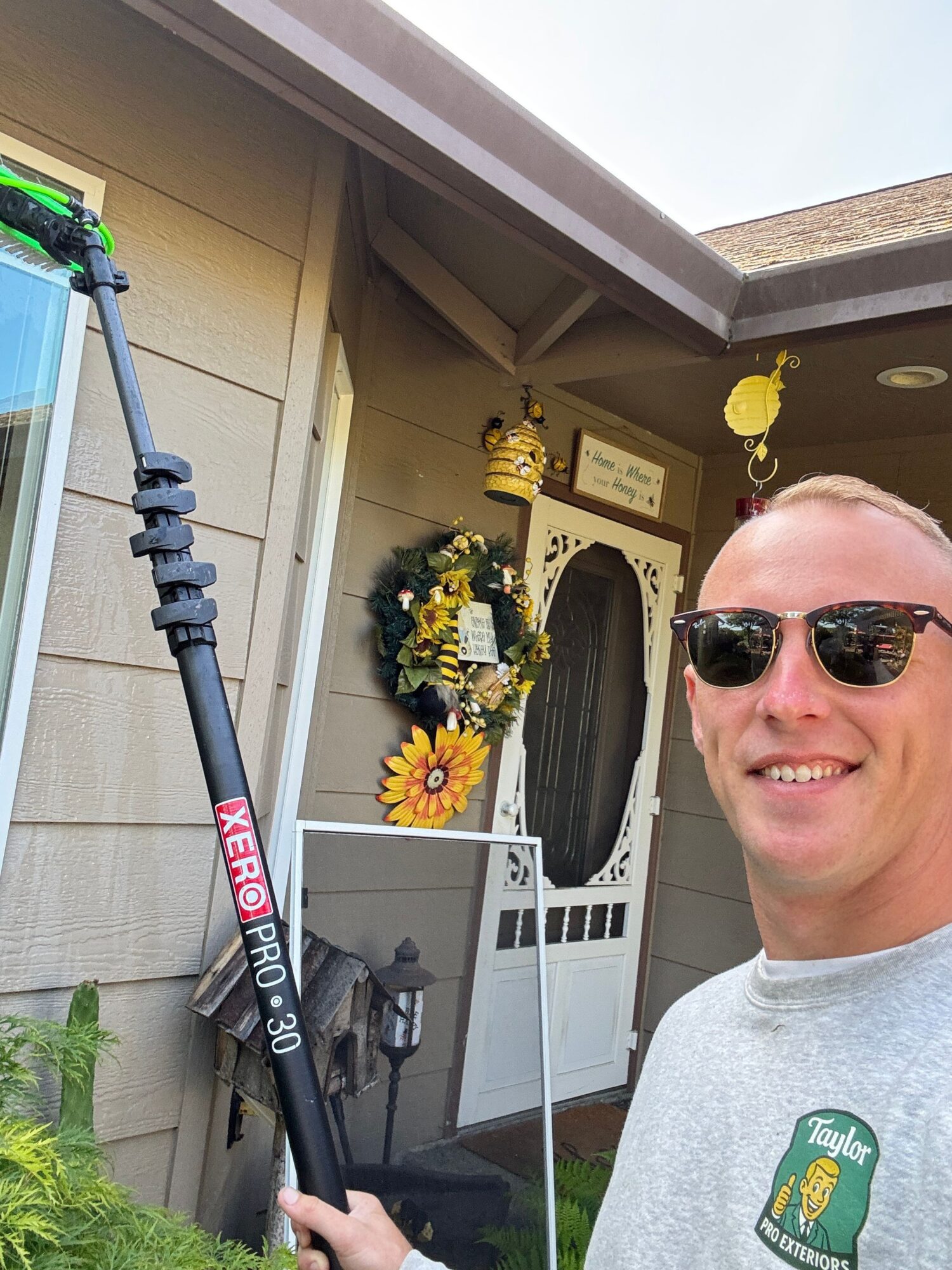 Smiling man wearing sunglasses and a gray shirt, standing outside a house with a door decorated with flowers and a wreath.