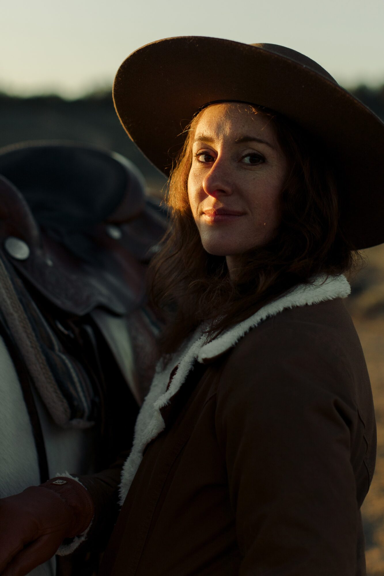 Woman wearing a large hat and coat outdoors, facing camera with a slight smile, sunset background.