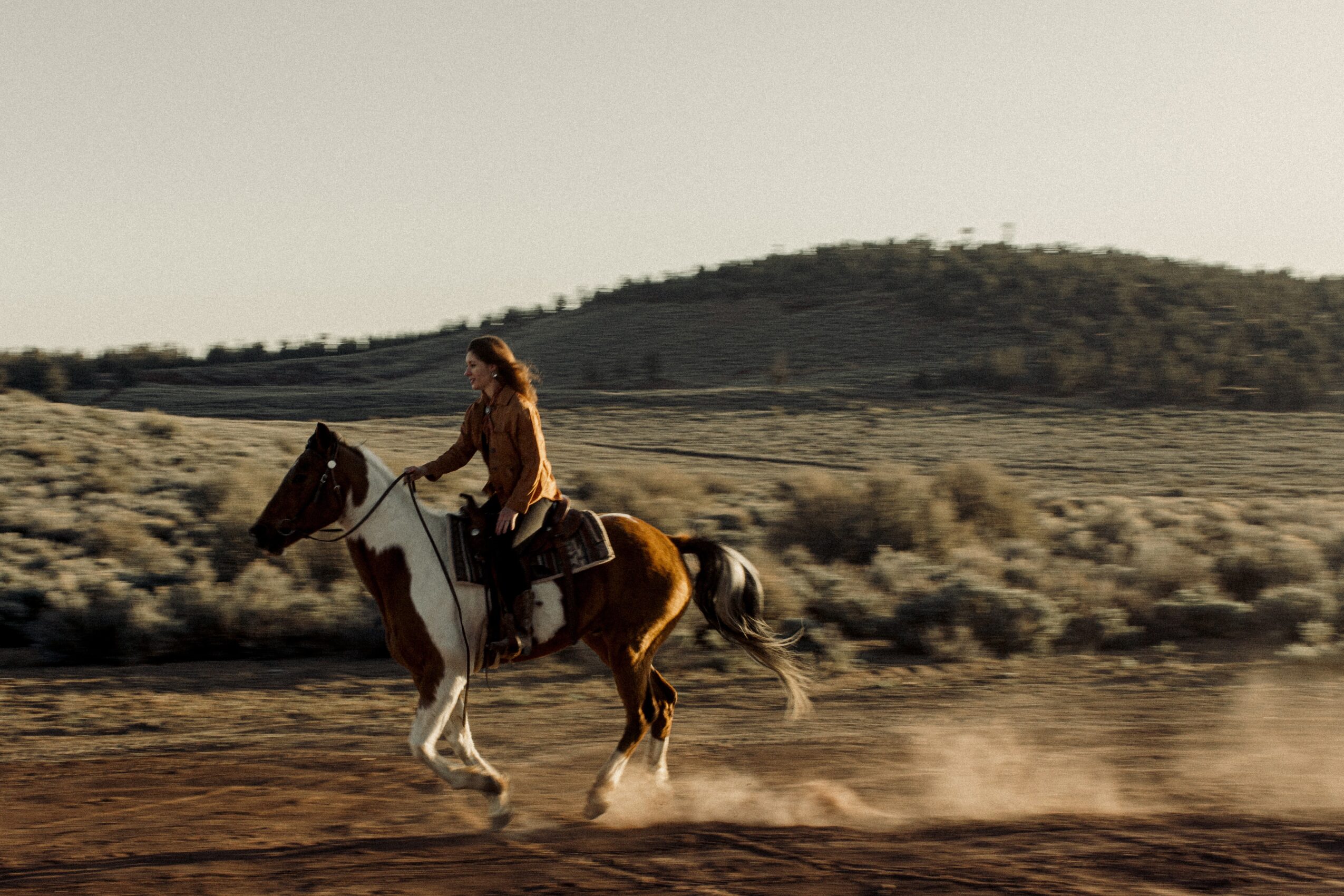 Person riding a horse on a dirt path with a hill in the background, dust rising from the horse's hooves.