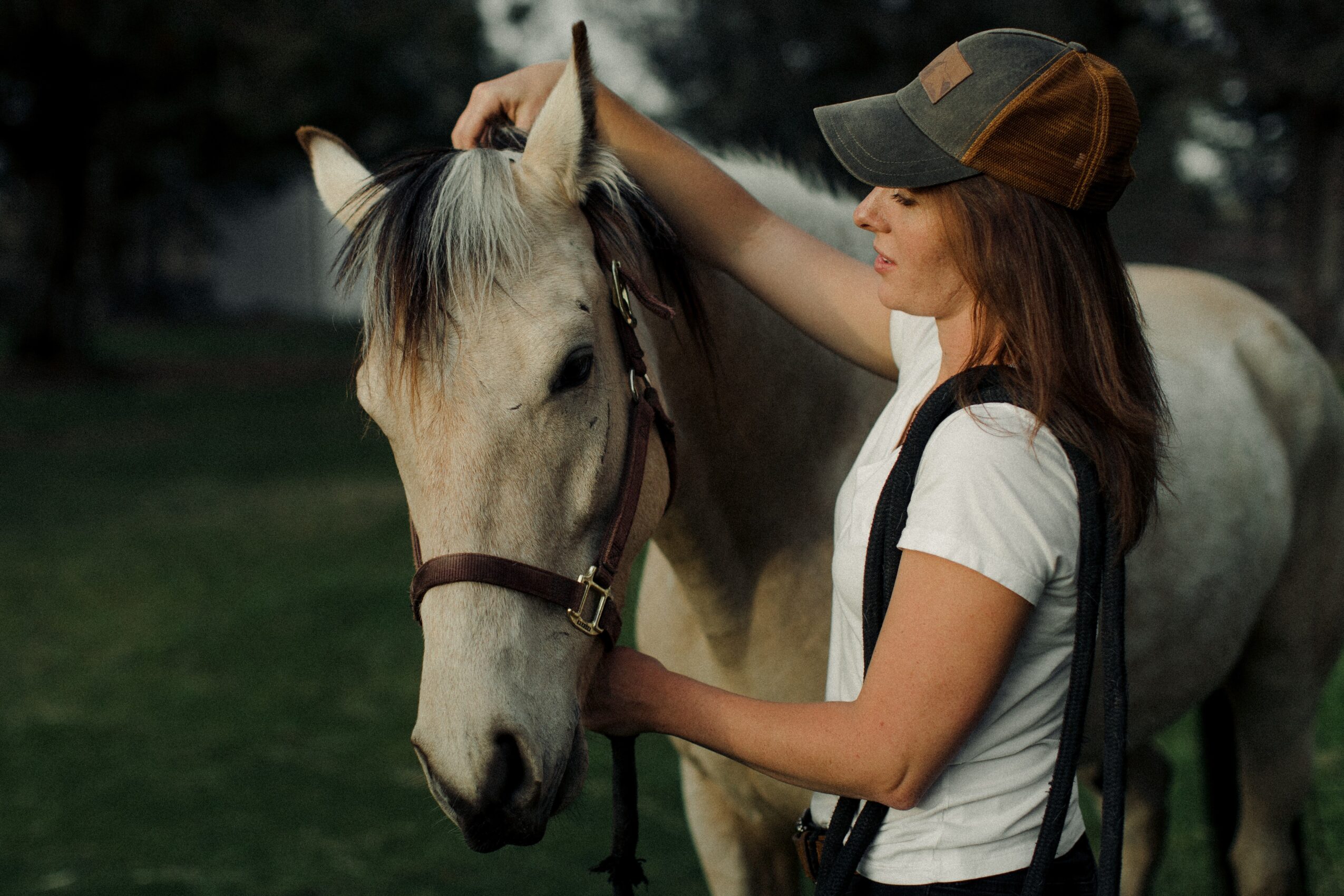 Woman petting a white horse outdoors during daytime, wearing a cap and casual clothing.