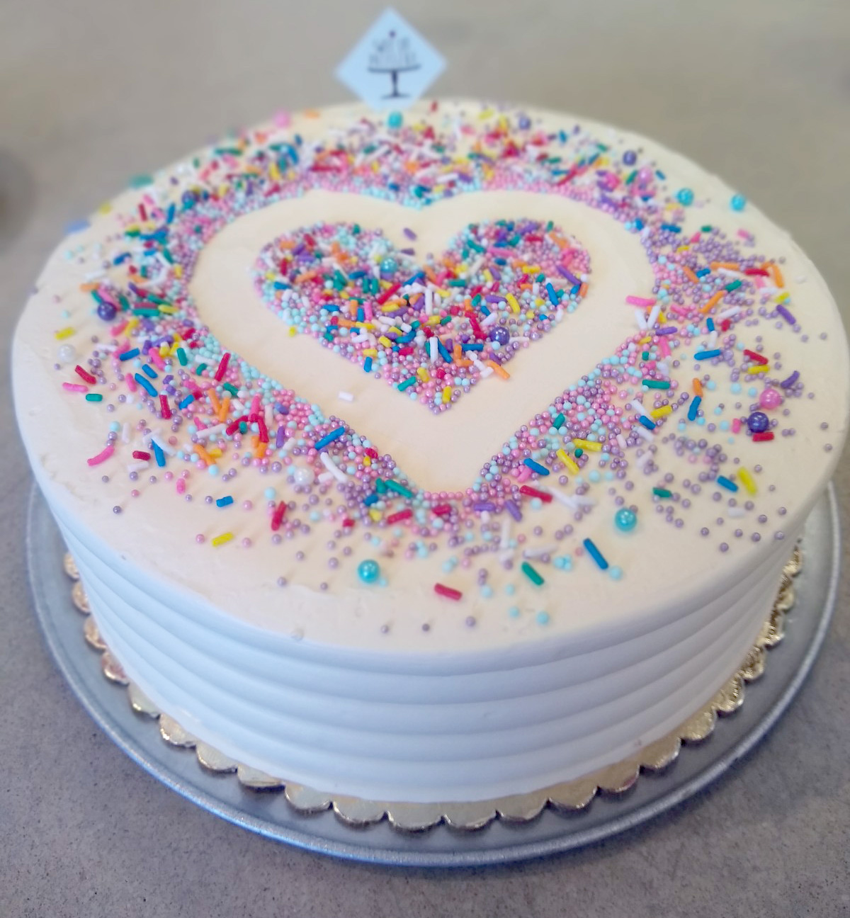 Round cake with white frosting, colorful sprinkles, and a heart shape in the center, on a silver base.