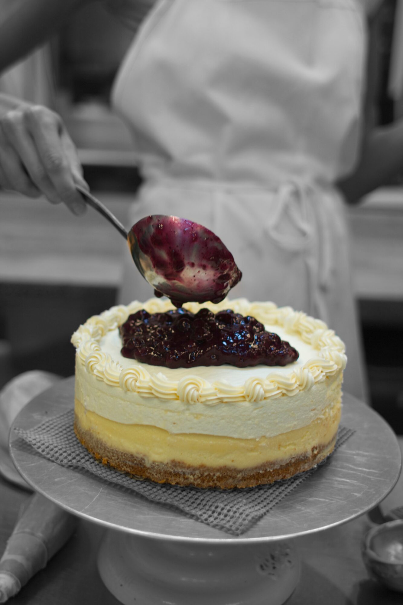Person in white apron adding fruit topping to a layered cake with white frosting, on a cake stand.
