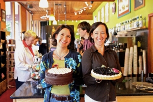 Two women smiling and holding cakes inside a cafe, with other customers in the background.