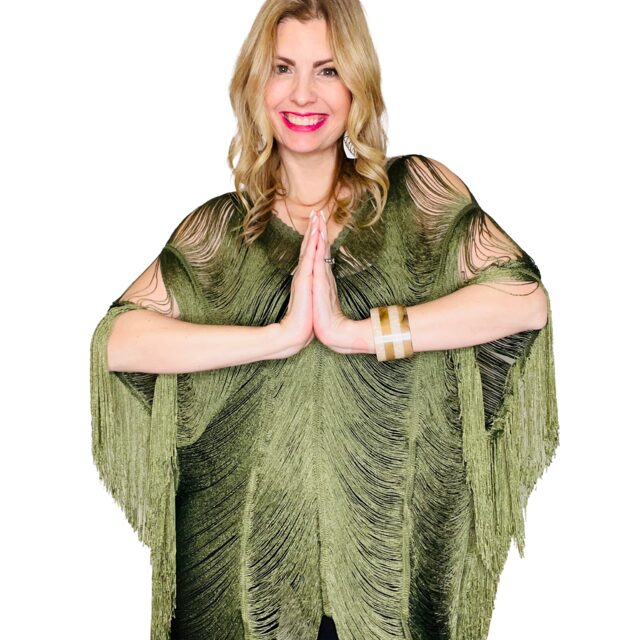 Woman with blonde hair smiling, hands pressed together in prayer, wearing a green fringed dress, against a white background.