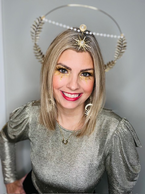 Woman with blonde hair smiling, wearing a metallic silver top and a decorative halo headpiece with gold accents.