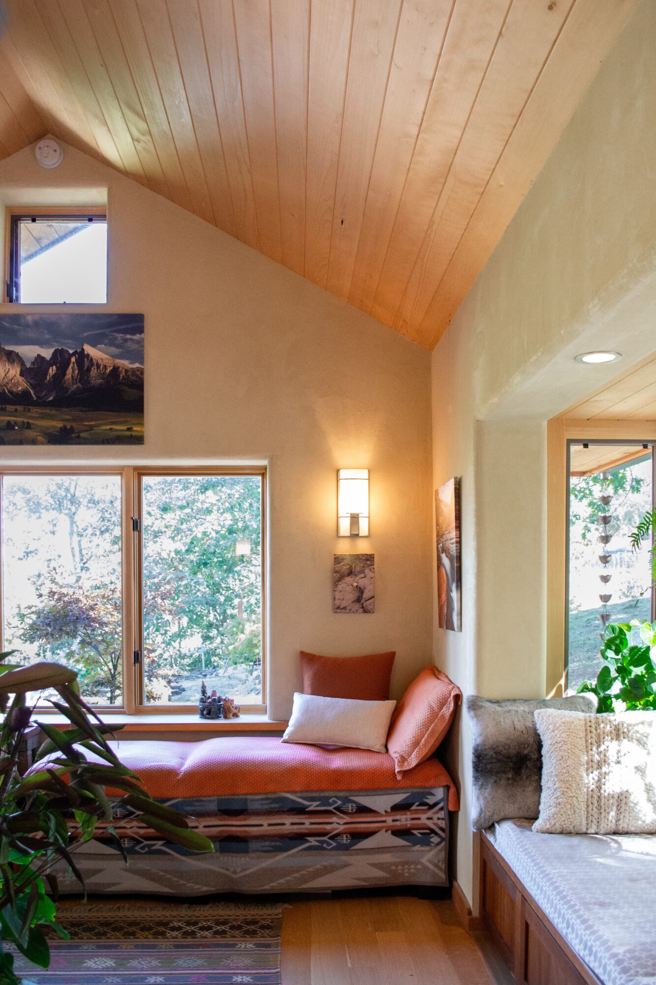 Living room with large window, wooden ceiling, pink sofa, and plants, bright natural light, and artwork on walls.