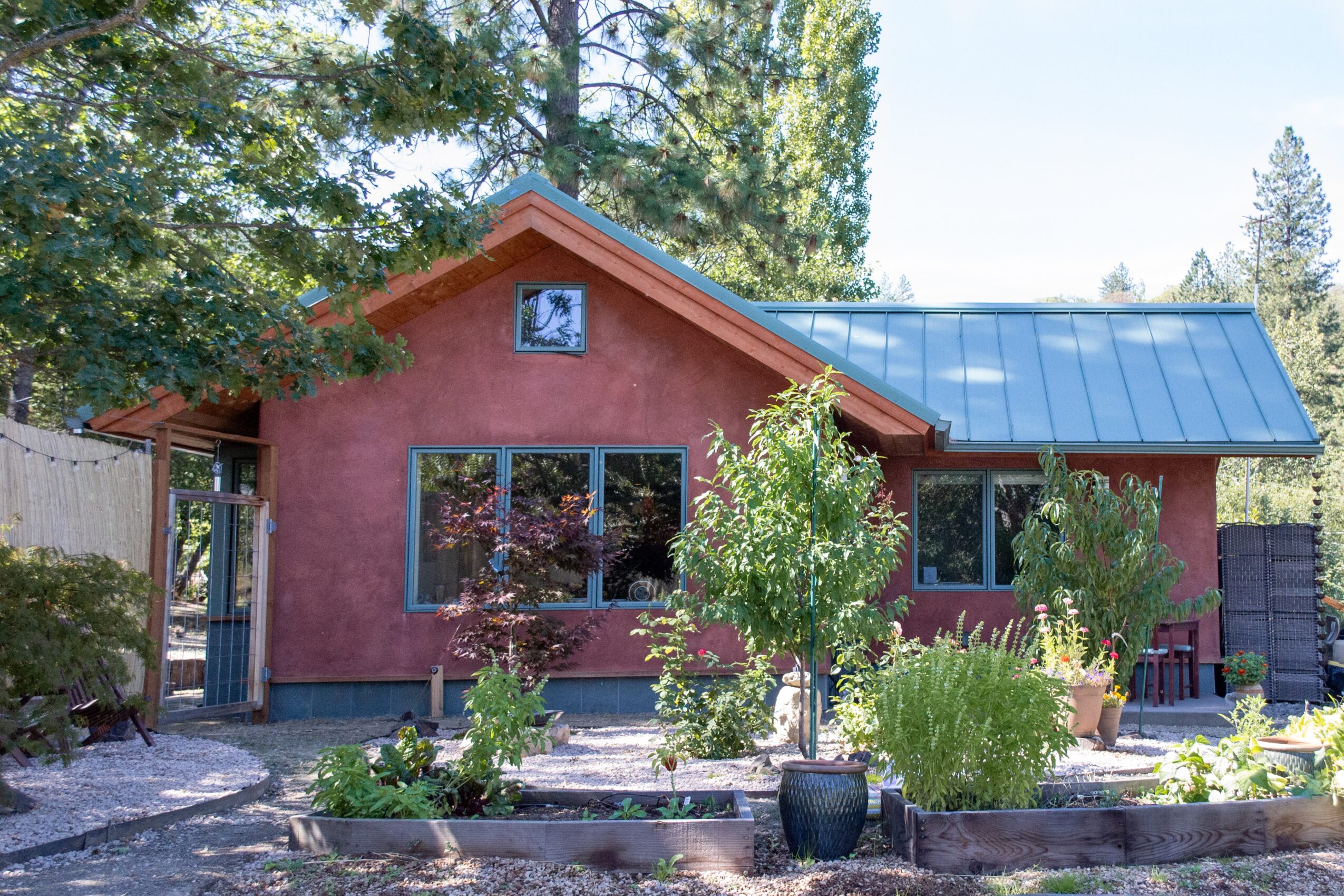 Small house with pink exterior, blue roof, surrounded by trees and plants, with a garden in front.