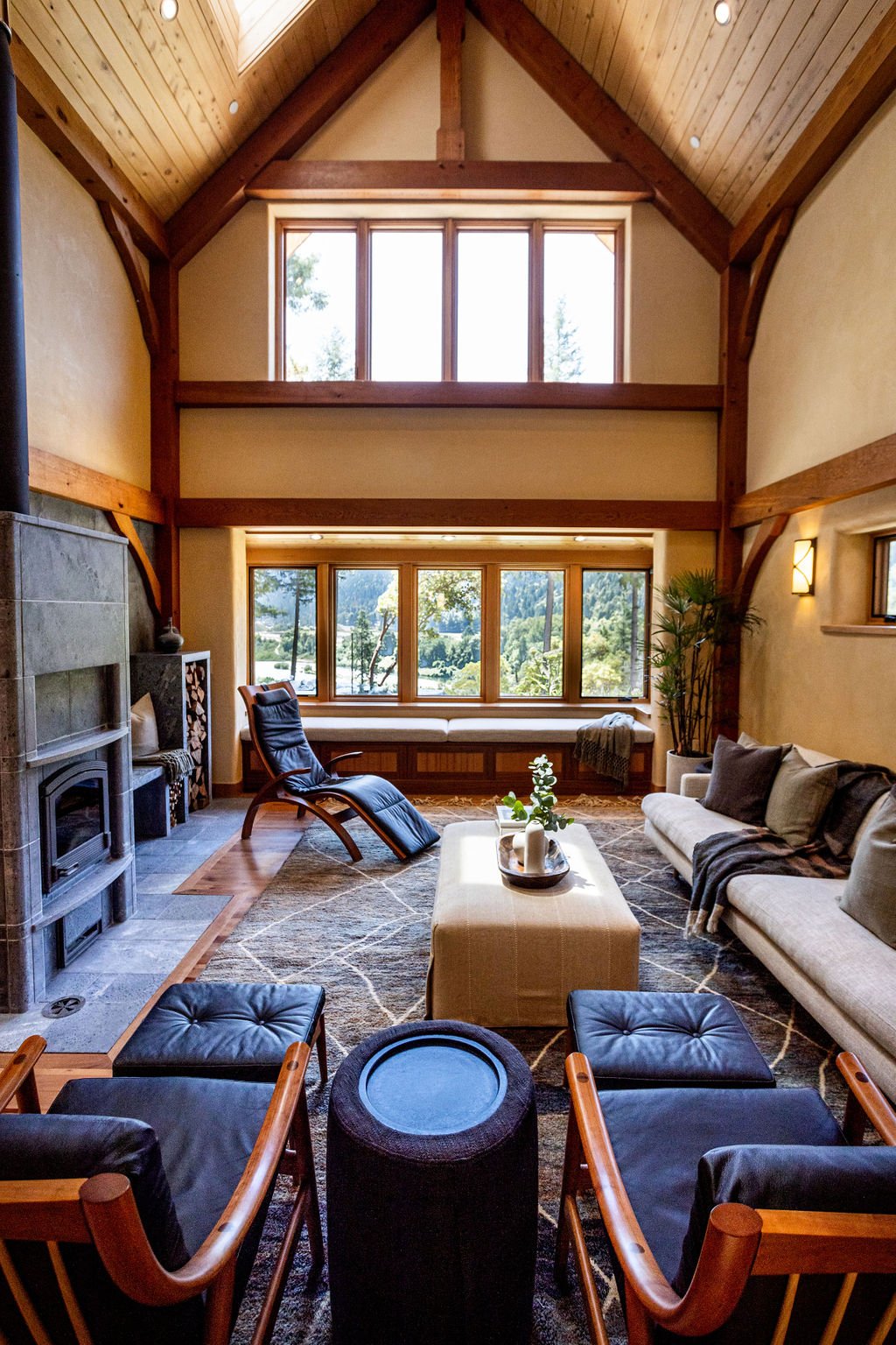 Living room with large windows, wooden ceiling, sofa, armchairs, and a coffee table, overlooking a scenic outdoor view.