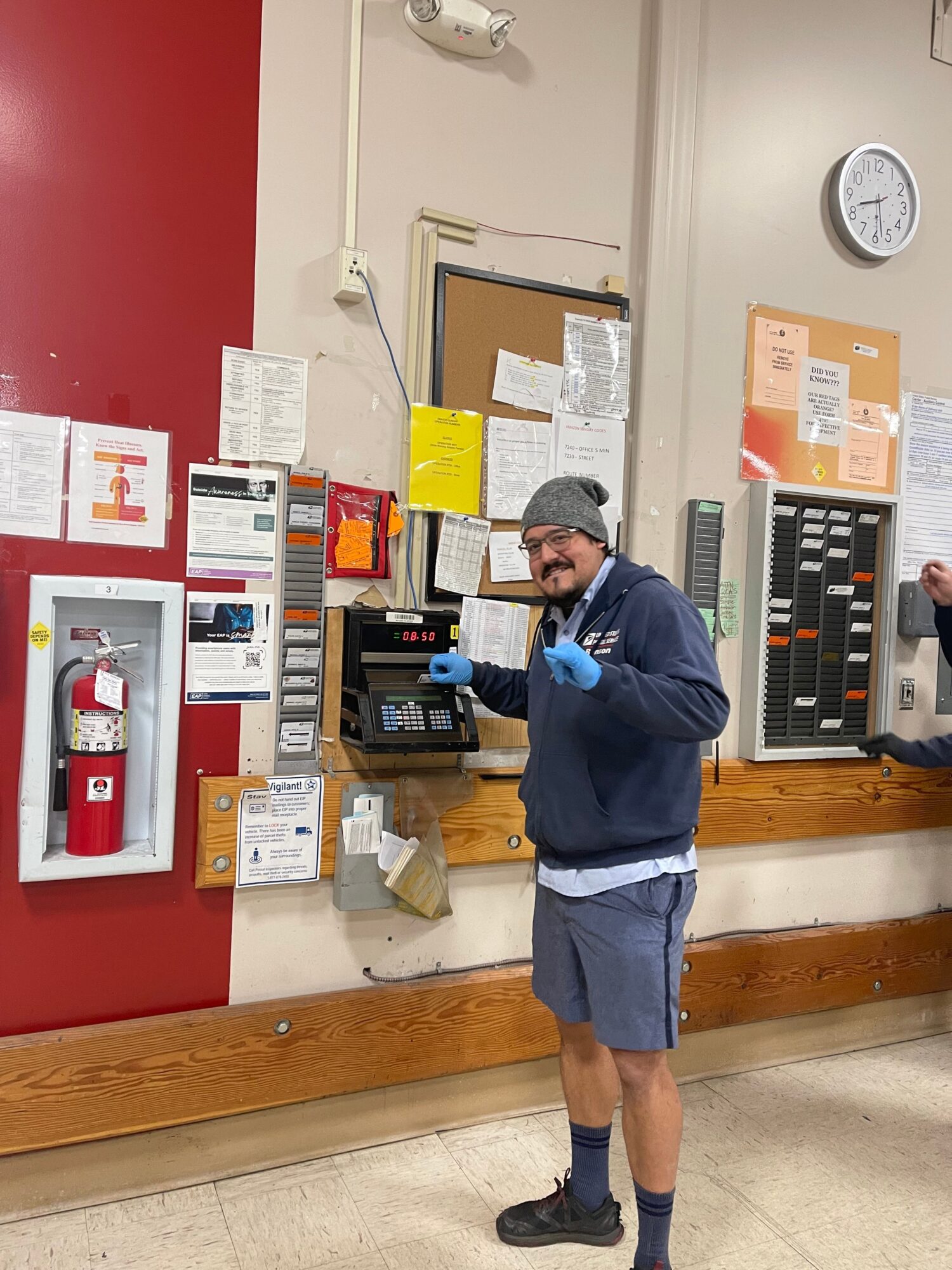 Child in a blue jacket and gray shorts stands in front of a vending machine, looking at it, with a person partially visible behind.