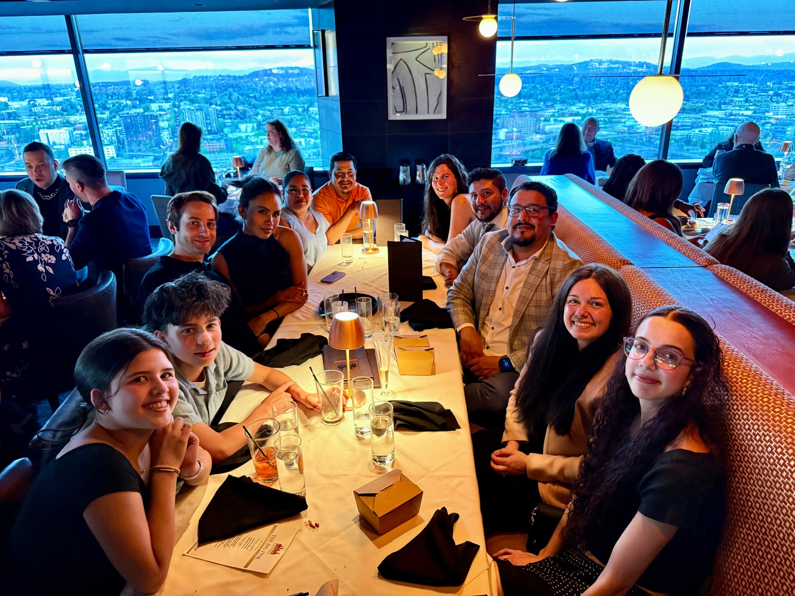 Group of people dining at a restaurant with large windows and city view, smiling and enjoying their meal.