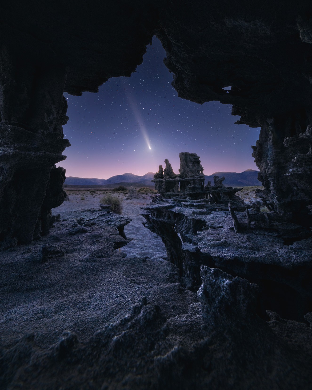 View from inside a cave looking out at a starry night sky with a bright streak, landscape, and rock formations.