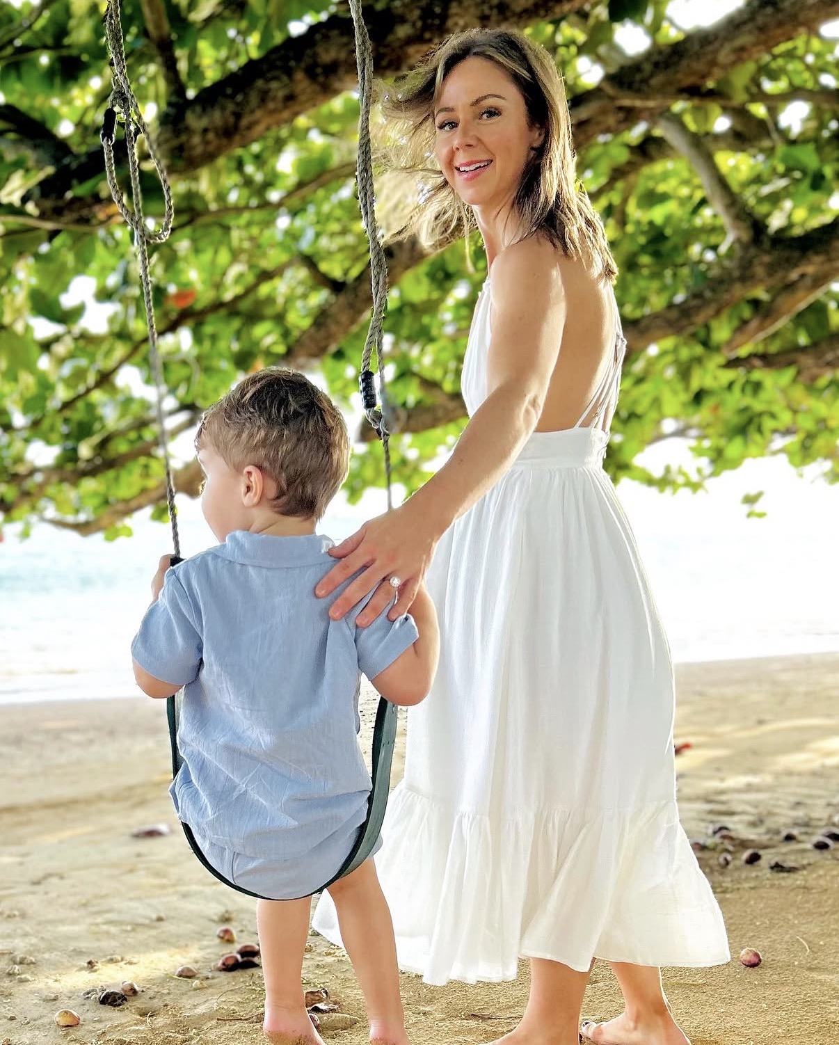Woman and child on a swing under a tree on a beach, woman smiling, child facing away, sunny day.