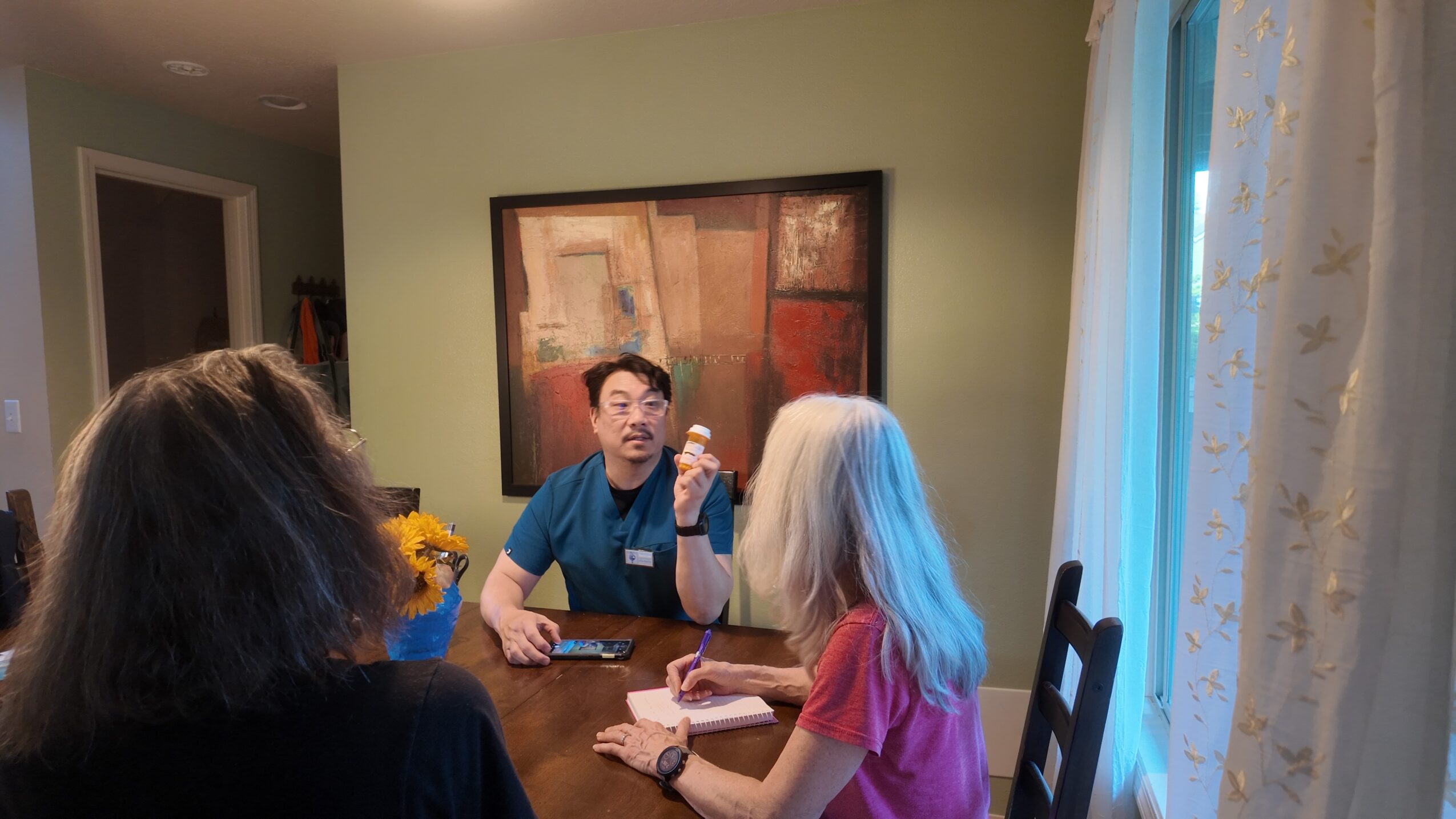 Three people sitting at a dining table in a room with a large window and artwork on the wall.