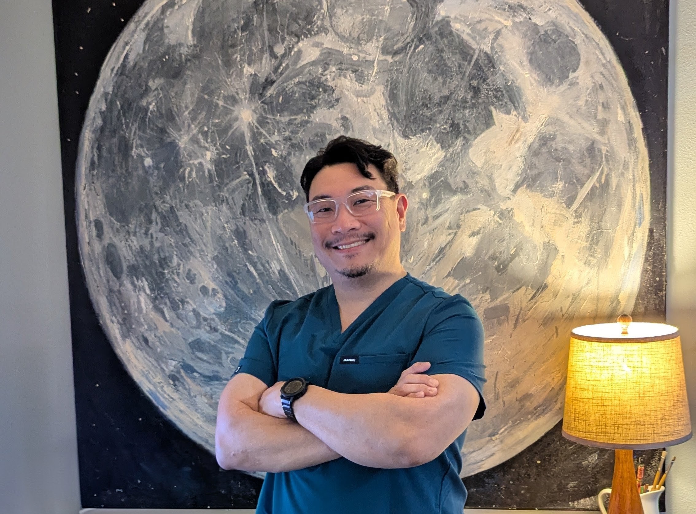 Man with glasses and a watch smiling with arms crossed in front of a large moon mural, standing next to a lamp.
