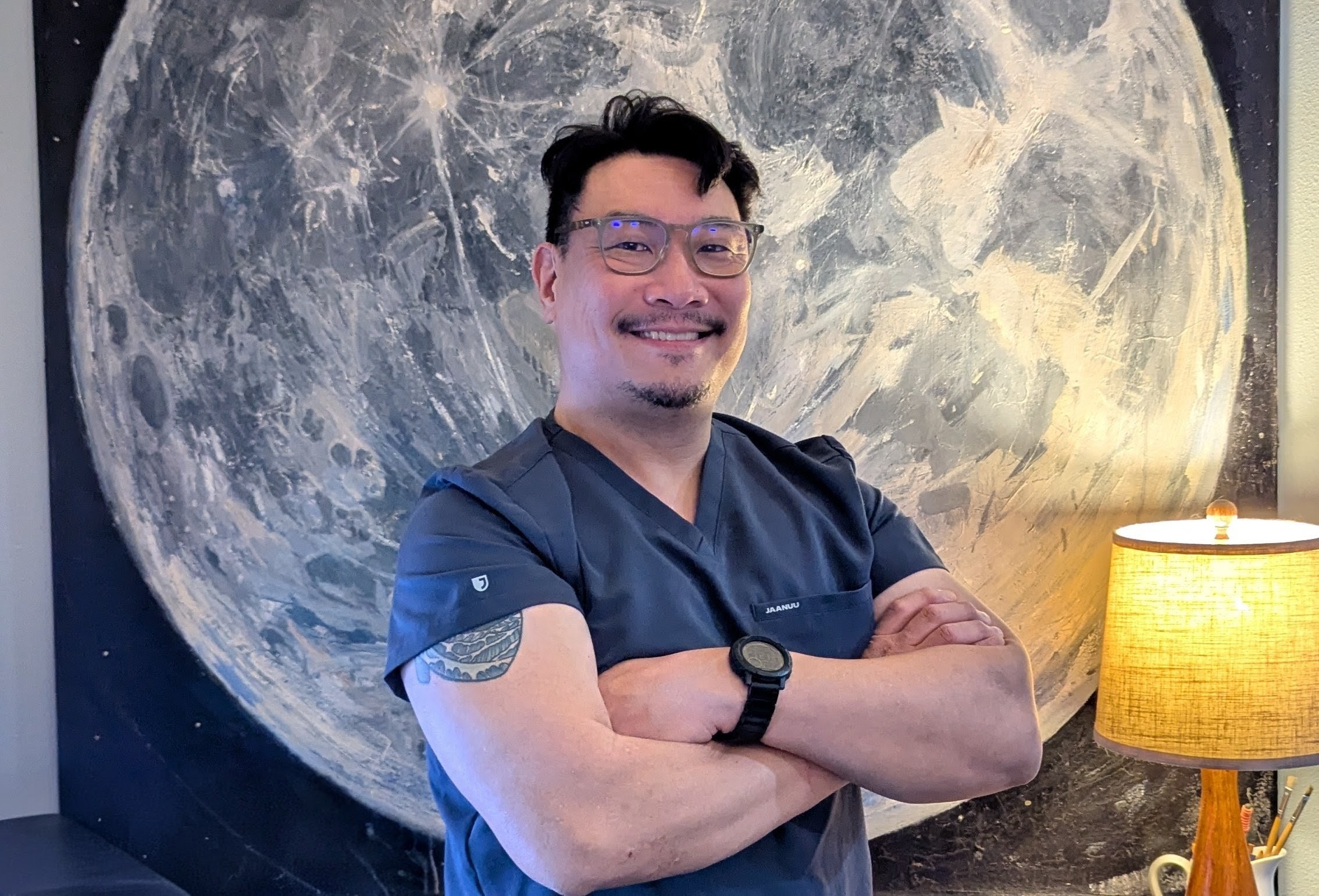 Man with glasses and a tattoo on his arm stands with arms crossed in front of a large moon mural, smiling.