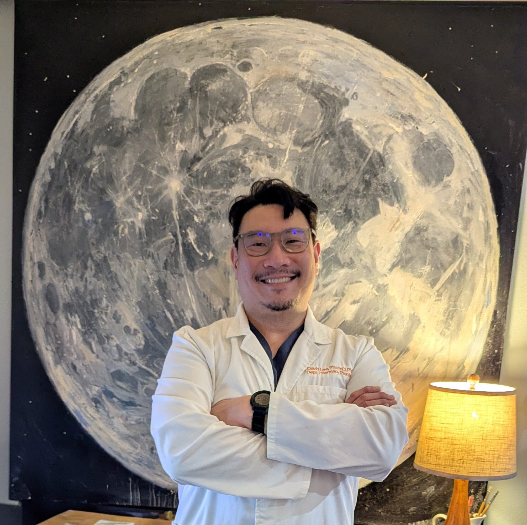 Man with glasses and a white coat smiling in front of a large moon mural, with a lamp nearby.
