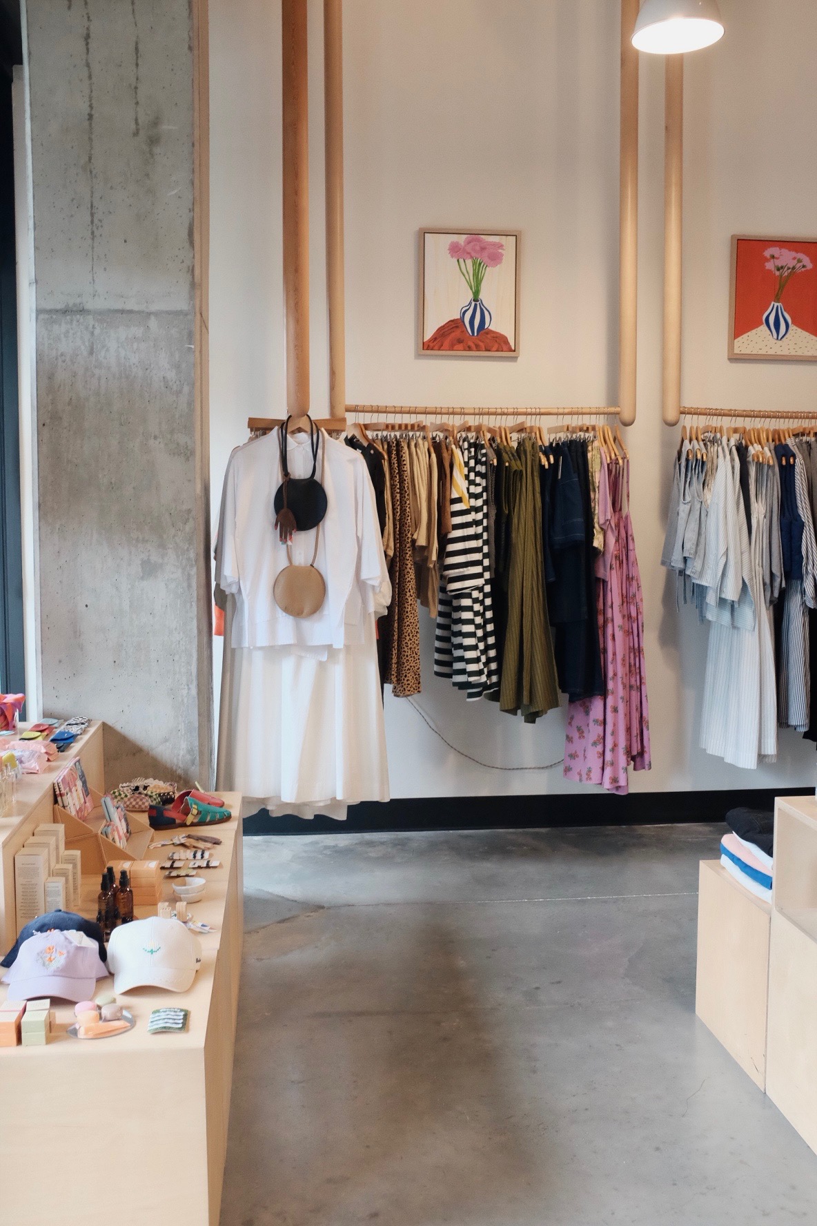 Clothing store interior with hanging dresses, skirts, and framed artwork on the wall, concrete floor, and display table.