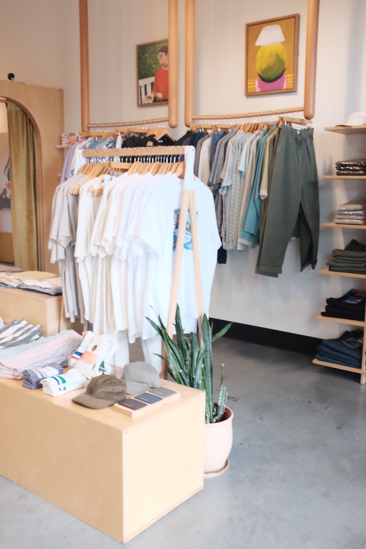 Clothing store with racks of hanging garments, a wooden display table, and framed artwork on the wall.