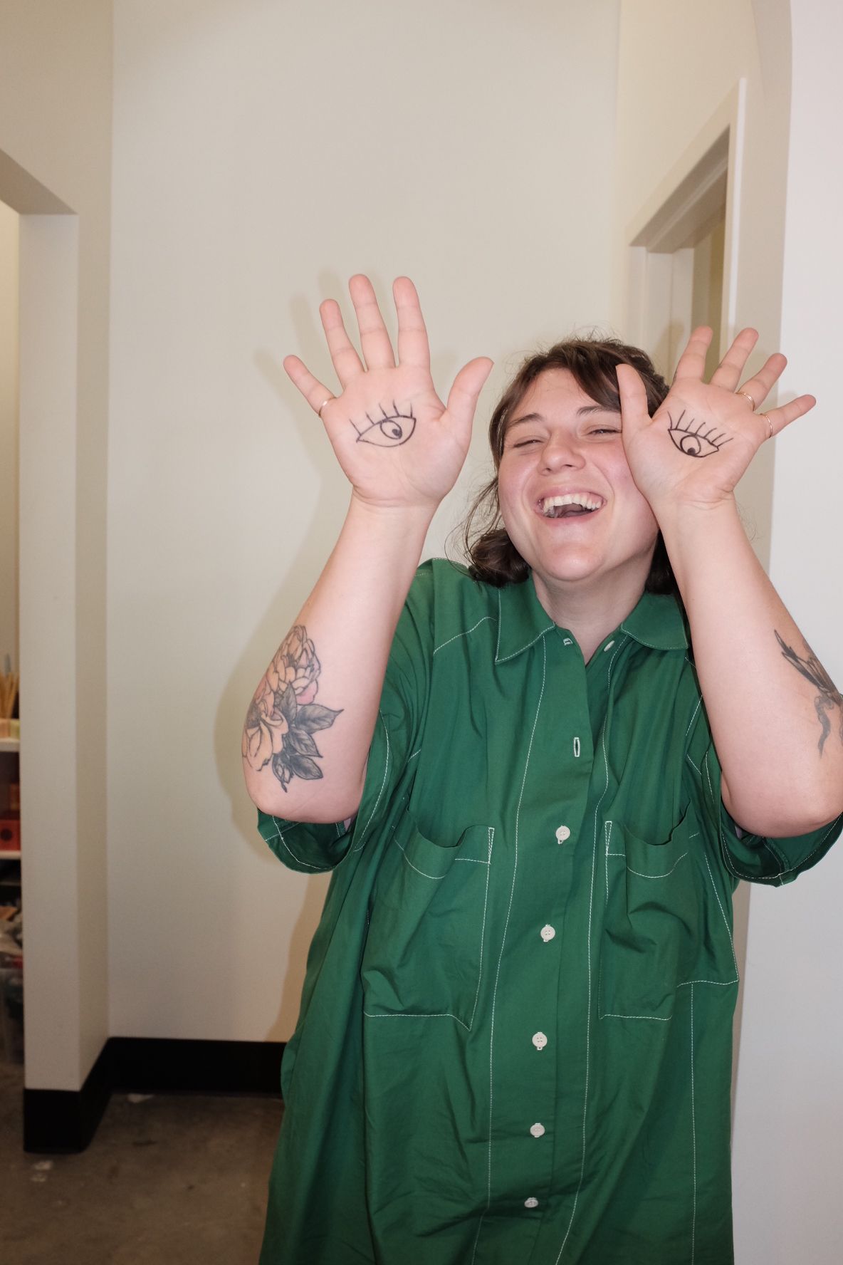 Person with short dark hair wearing a green shirt, smiling, holding up hands with eye drawings, standing indoors.