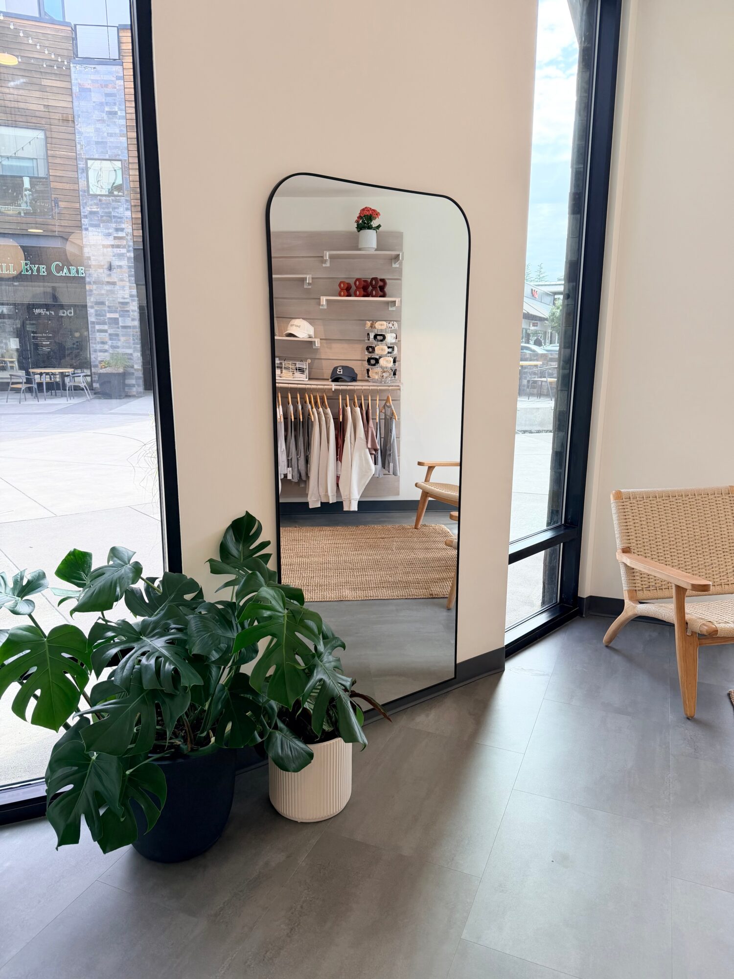 Full-length mirror reflecting a clothing display and shelves inside a store, with a plant and chair nearby.